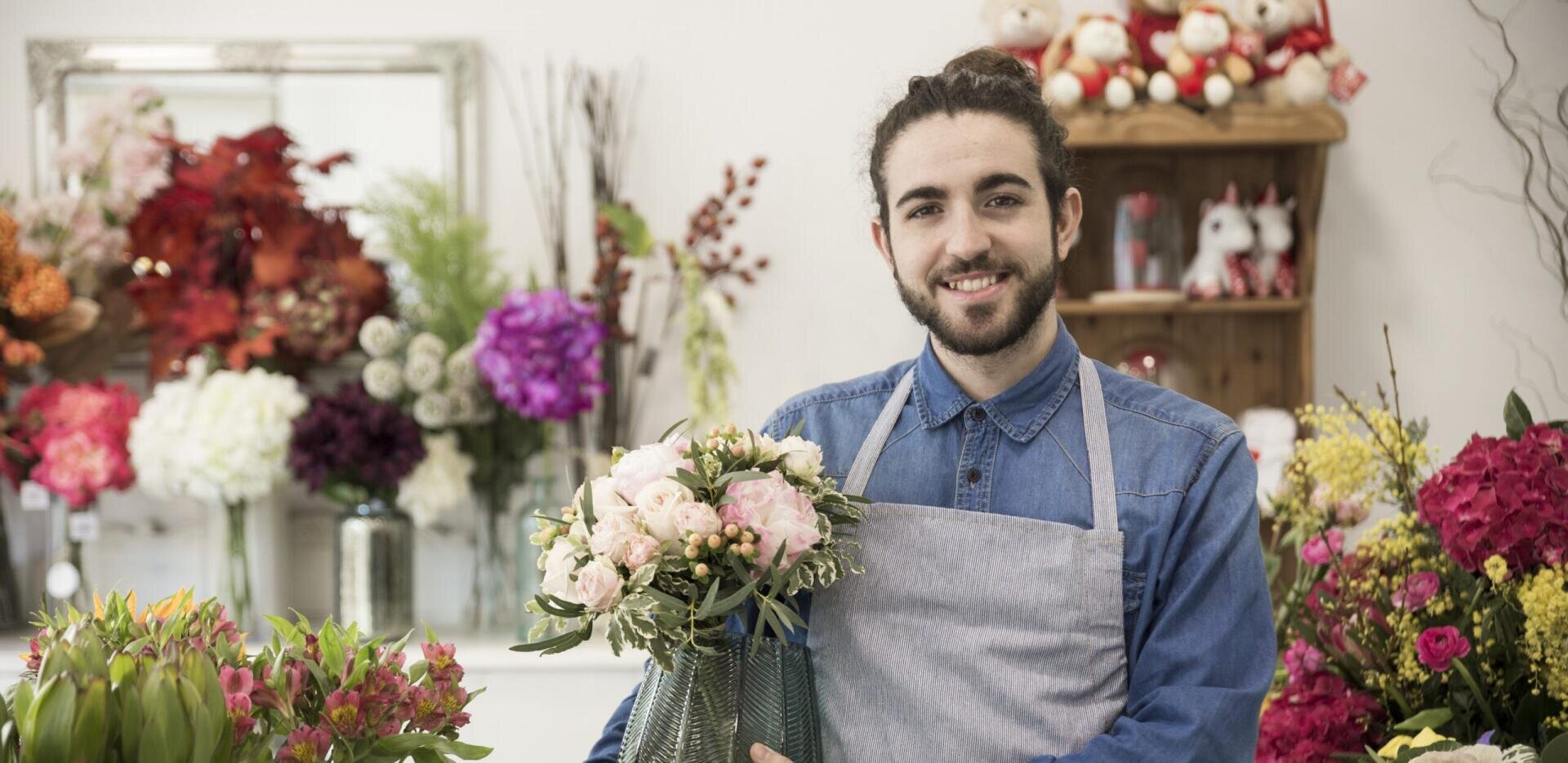 Ein lächelnder Florist in einem Jeanshemd und einer Schürze hält einen Strauß aus rosa und weißen Blumen in der Hand, umgeben von bunten Blumenarrangements in einem Geschäft.