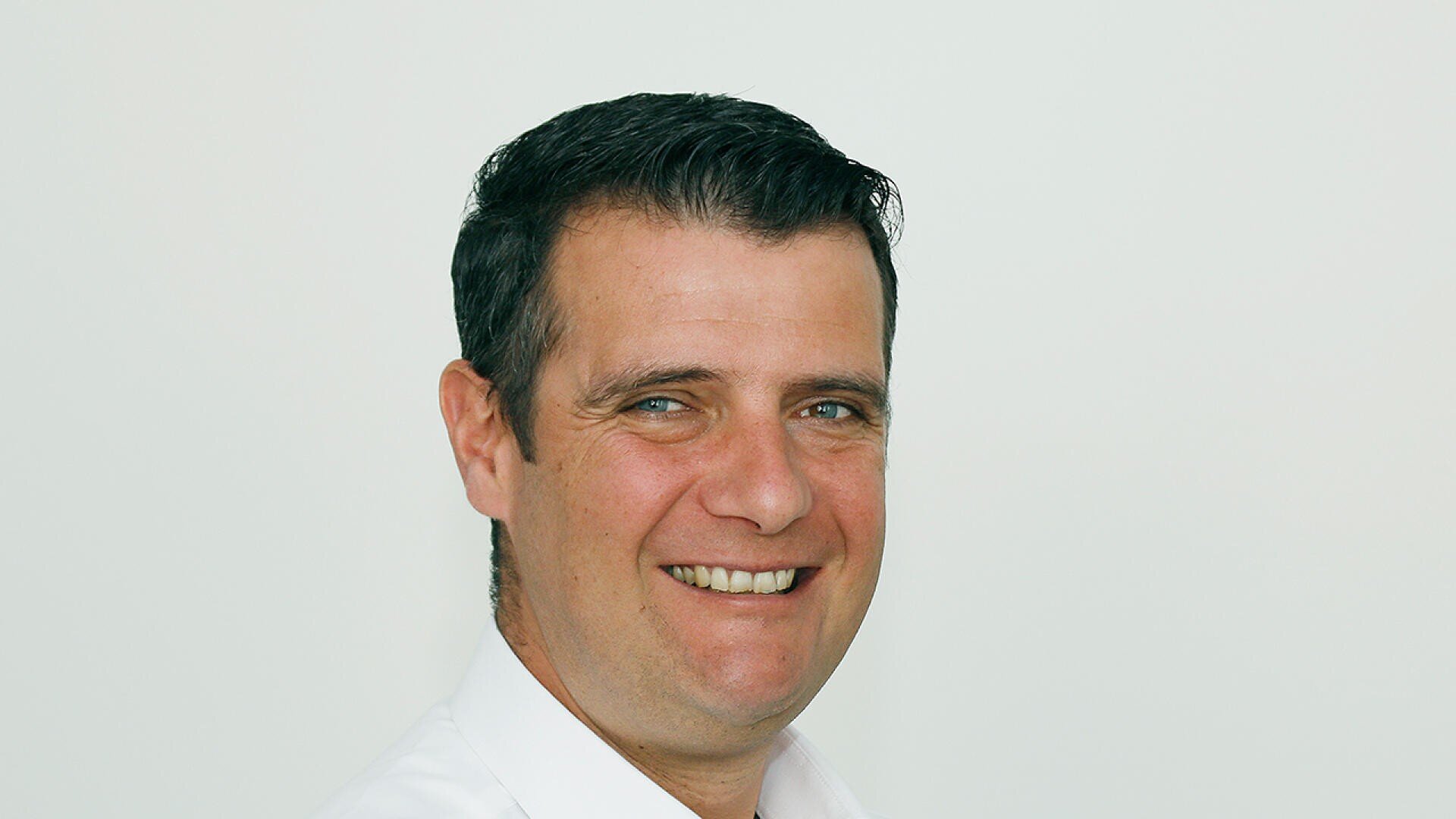 Ein lächelnder Mann mit kurzen dunklen Haaren, der ein weißes Hemd trägt und vor einem schlichten, hellen Hintergrund in die Kamera blickt.