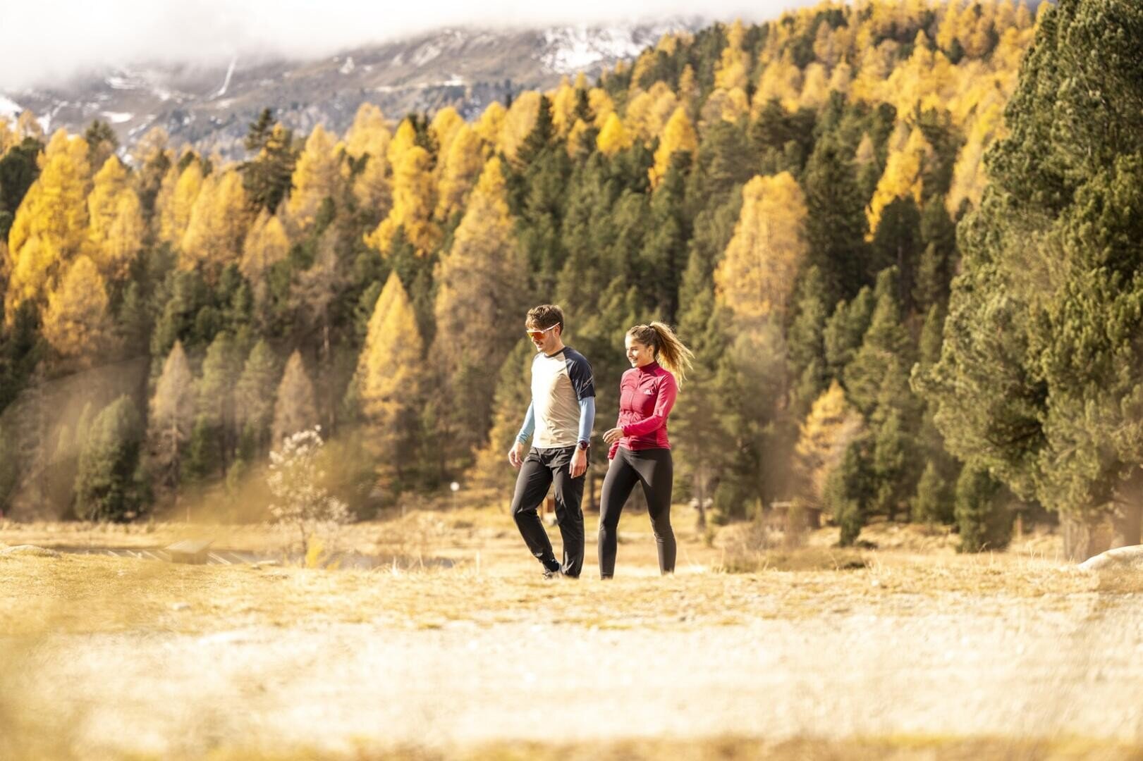Zwei Tourismusstudierende der HFT Graubünden spazieren gemeinsam über eine Wiese im Engadin, im Hintergrund sind herbstlich gefärbte Bäume und Berge zu sehen, und genießen einen sonnigen Tag im Freien.