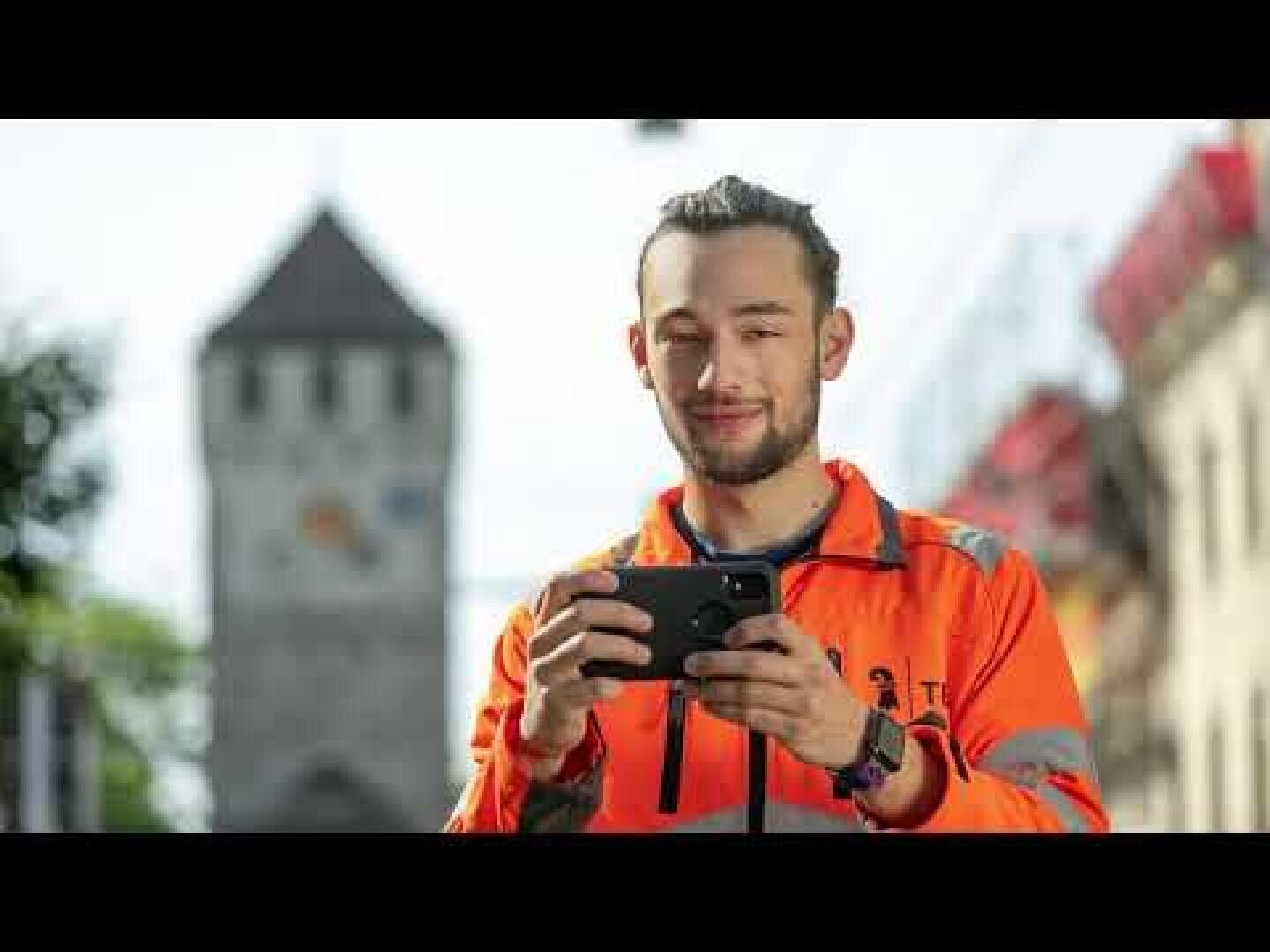 Ein Mann in einer orangefarbenen Warnweste hält ein Smartphone in der Hand und lächelt im Freien, im Hintergrund sind ein historischer Uhrturm und Gebäude zu sehen.