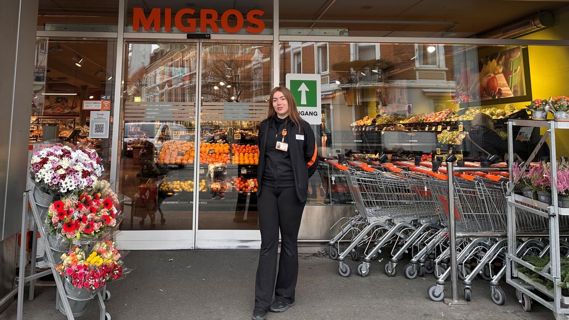 Eine Frau in schwarzer Kleidung steht vor dem Eingang einer Migros-Filiale, in der Nähe von Blumenauslagen und Einkaufswagen; im Inneren sind Obst und Gemüse zu sehen.