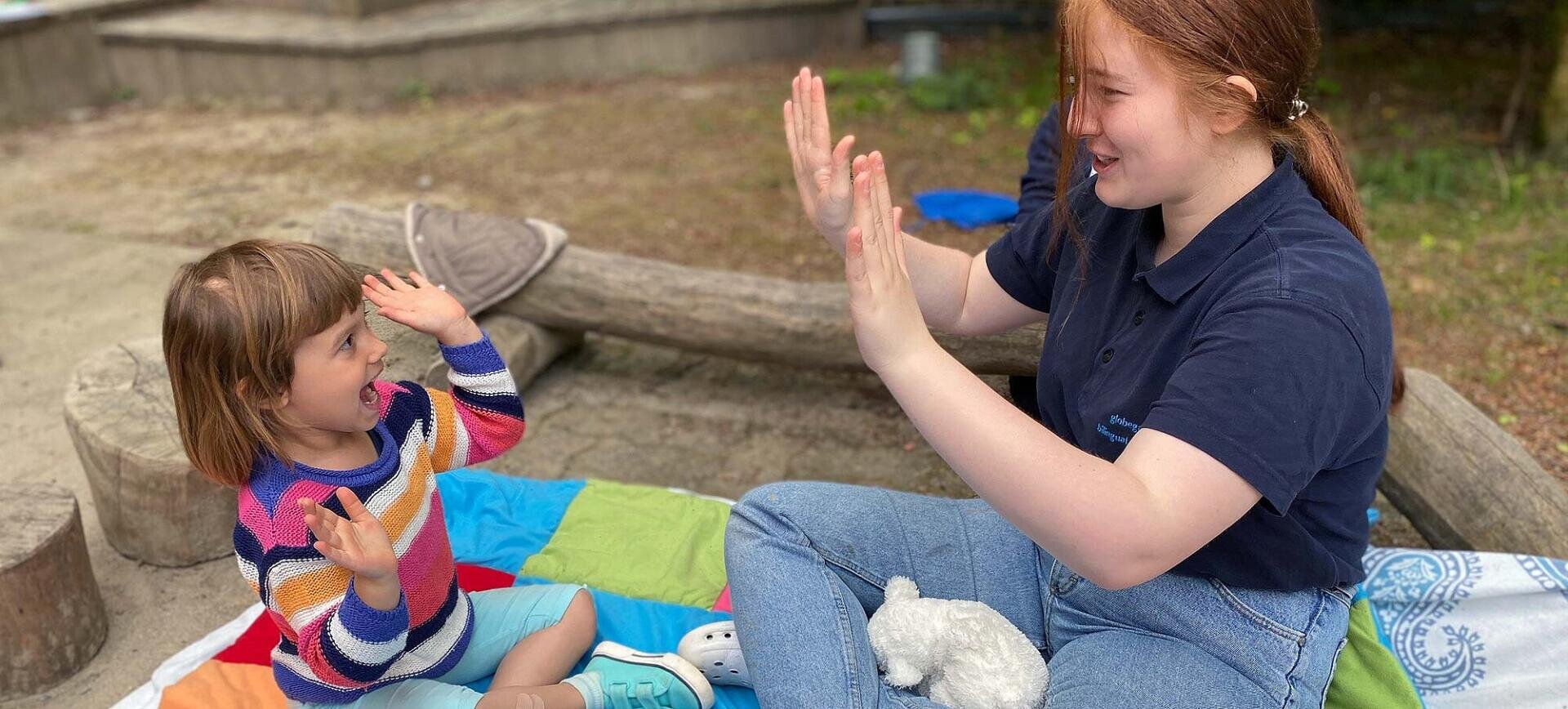 Eine junge Frau und ein Kind sitzen auf einer farbenfrohen Decke im Freien, lächeln und spielen zusammen ein Handklatschspiel. Das Kind trägt einen gestreiften Pullover und hat einen Gips an einem Bein. Im Hintergrund sind Baumstämme und Gras zu sehen.