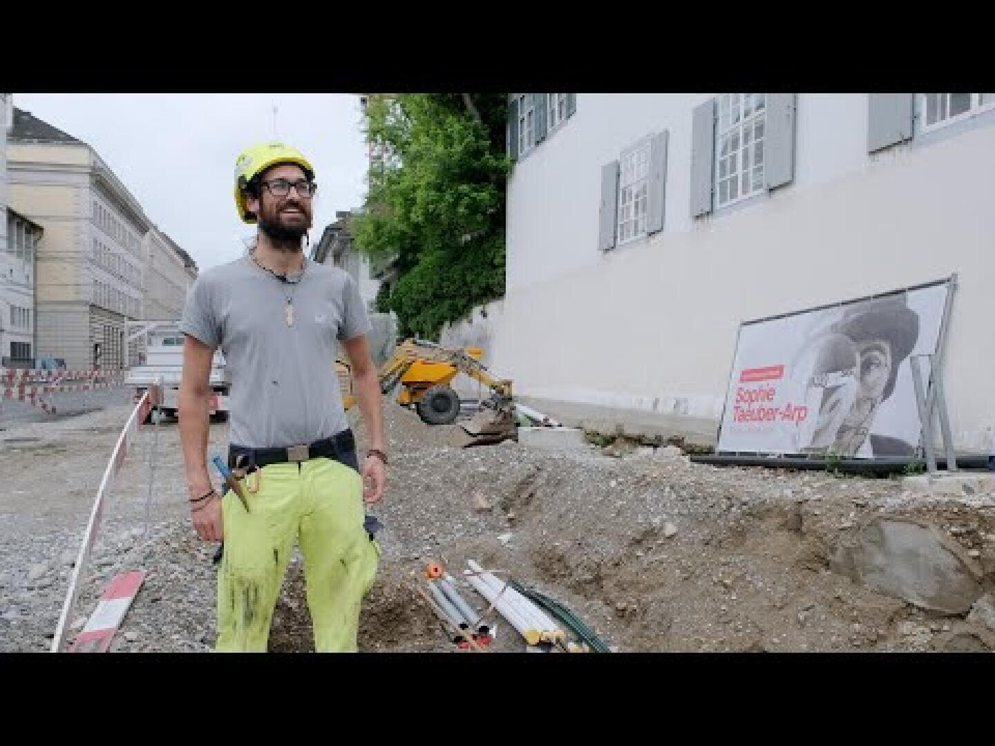 Ein Bauarbeiter in einem grauen Hemd und einem gelben Schutzhelm steht auf einer Baustelle, auf der sich Bauschutt, Werkzeuge und eine kleine Maschine befinden. Im Hintergrund sind ein großes Plakat und Gebäudefassaden zu sehen.