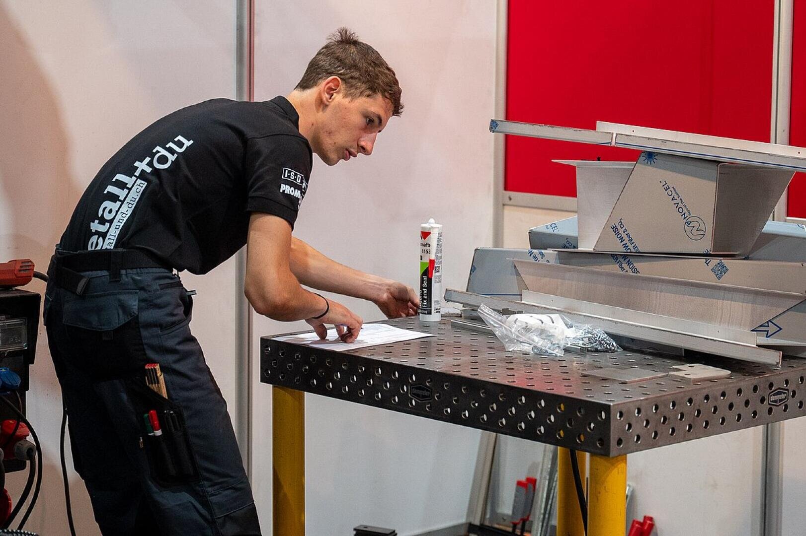 Ein junger Mann in einem schwarzen Hemd arbeitet an einem Metalltisch, liest Unterlagen und ist von Metallteilen und Werkzeugen umgeben. Auf dem Tisch steht eine Tube Dichtungsmasse. Im Hintergrund sind rote und weiße Paneele zu sehen.