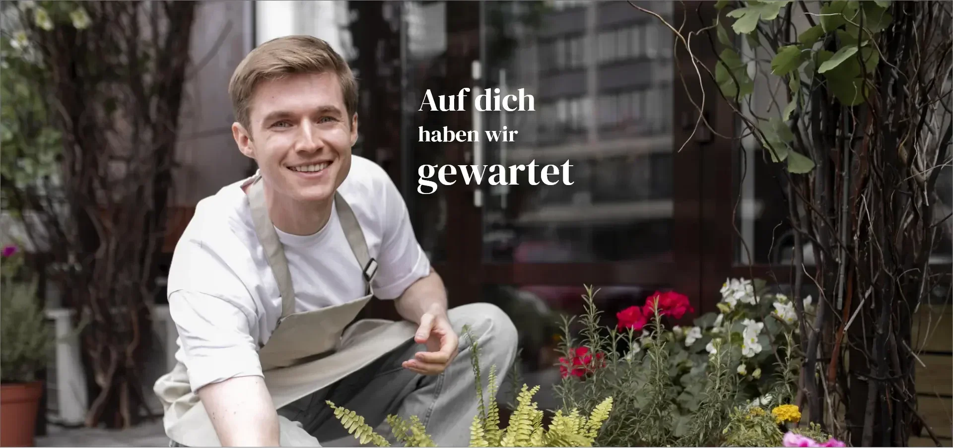 Eine lächelnde Person in einer Schürze hockt neben Pflanzen vor einem Laden, auf dessen Schild Auf dich haben wir gewartet steht.