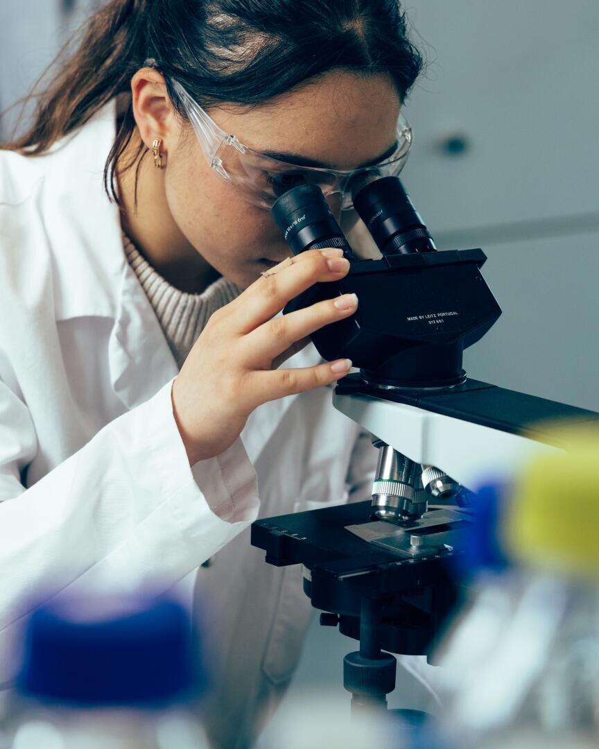 Eine Person in einem Laborkittel und mit Schutzbrille blickt durch ein Mikroskop und konzentriert sich dabei auf eine Probe in einer Laborumgebung. Im Vordergrund sind Flaschen und wissenschaftliche Geräte zu sehen.