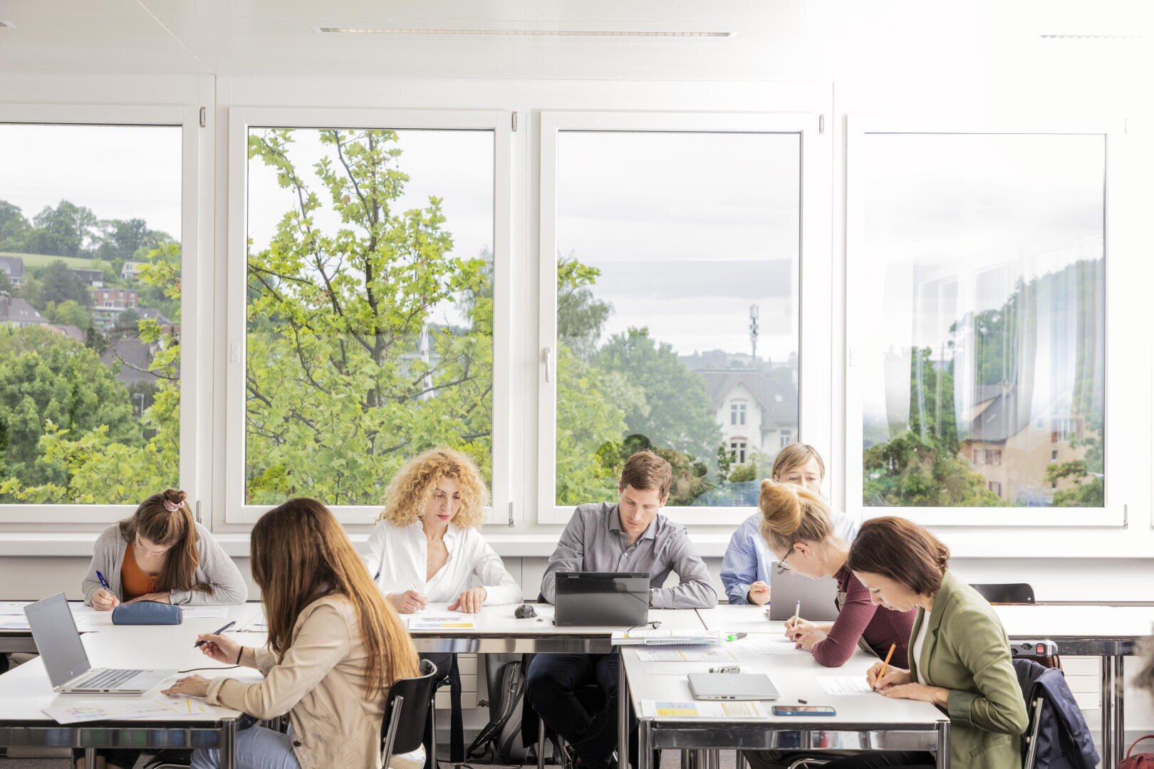 Sieben Personen sitzen an Schreibtischen in der Nähe großer Fenster, arbeiten an ihren Laptops, schreiben und lesen Unterlagen. Helles Tageslicht durchflutet den Raum, und draußen sind grüne Bäume und Gebäude zu sehen. Die Kulisse scheint ein modernes Klassenzimmer oder Büro zu sein.