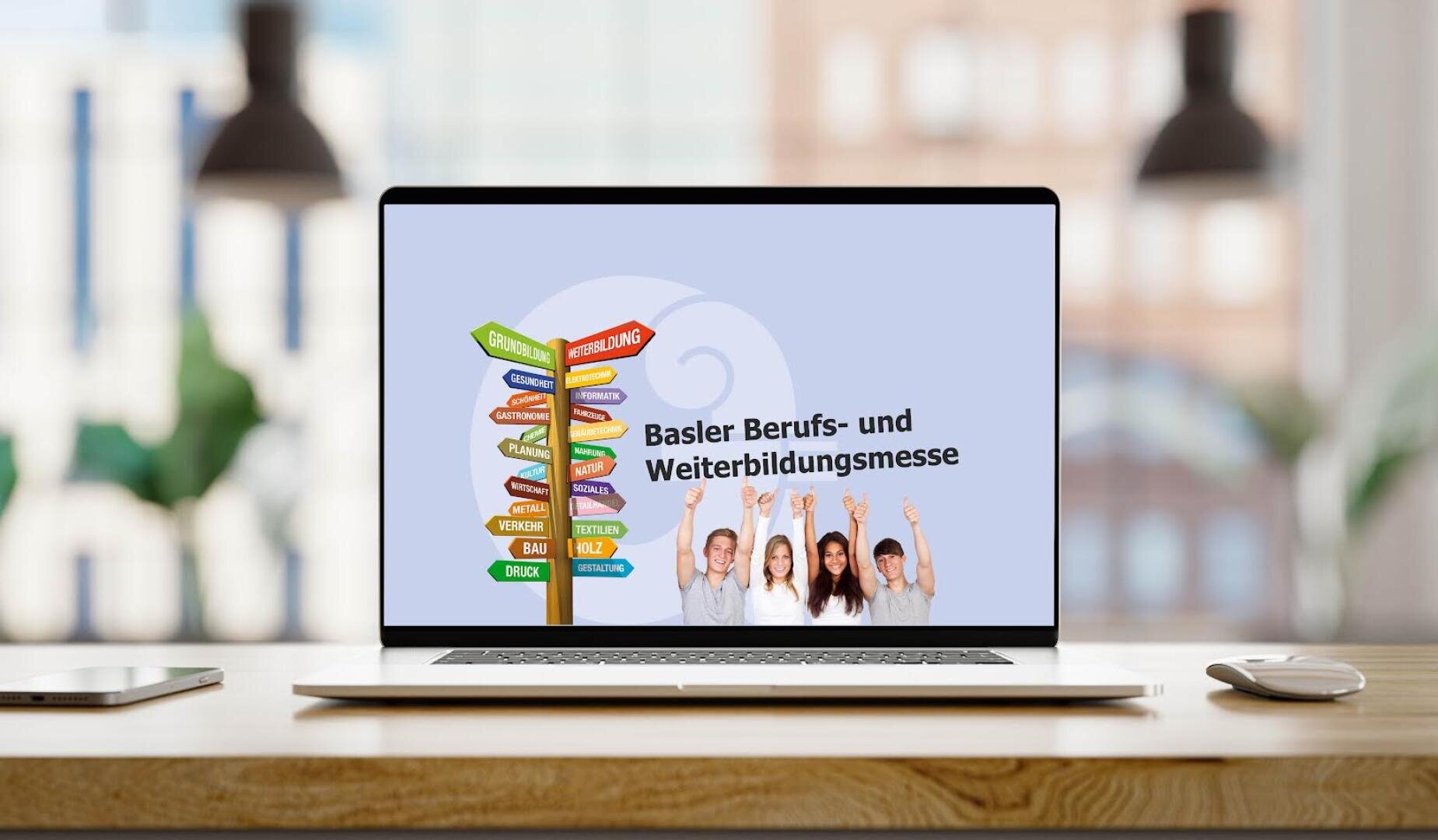 Auf einem Laptop auf einem Schreibtisch ist auf dem Bildschirm ein bunter Wegweiser, lächelnde Menschen, die ihre Hände heben, und der Text „Basler Berufs- und Weiterbildungsmesse“ auf hellem Hintergrund zu sehen.