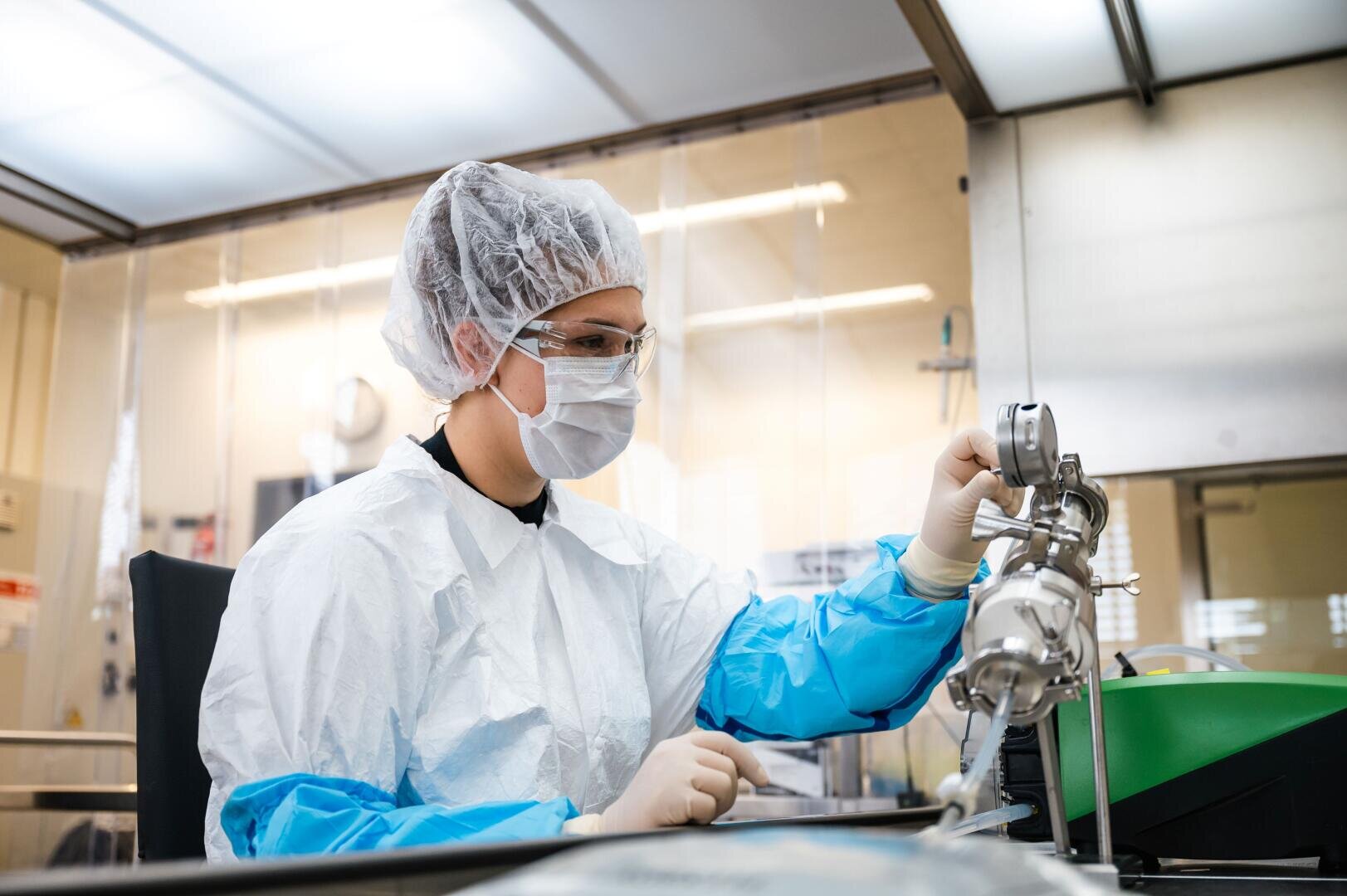 Ein Wissenschaftler, der Schutzkleidung, ein Haarnetz, eine Maske und eine Schutzbrille trägt, bedient Laborgeräte in einer sauberen, sterilen Laborumgebung.