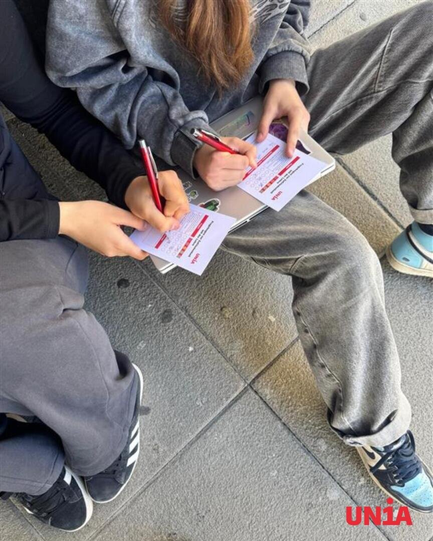 Zwei Personen sitzen auf einem Bürgersteig und schreiben mit Stiften auf kleine weiße und rote Kärtchen. Eine Person hält einen Laptop in der Hand. Beide tragen legere graue Kleidung und Turnschuhe. In der rechten unteren Ecke ist das Wort „UNJA“ zu sehen.