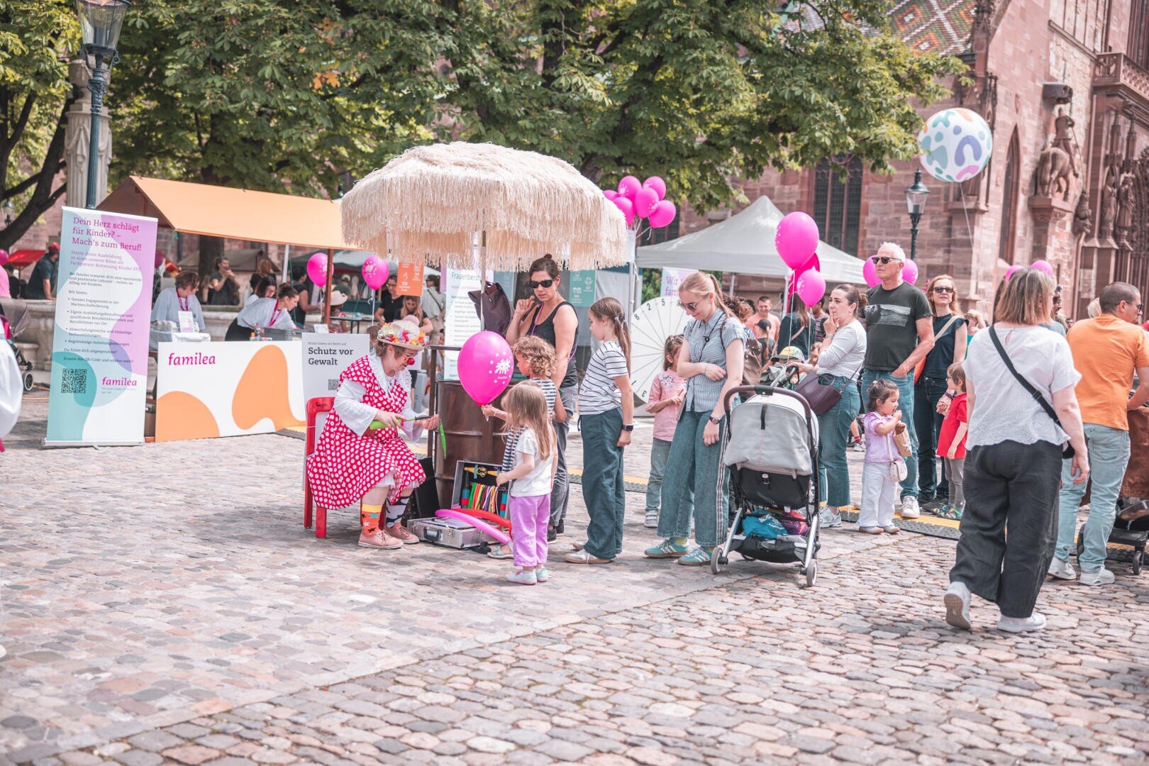 Eine Frau in einem roten Kleid mit Polka-Punkten sitzt unter einem Sonnenschirm und bastelt für Kinder Ballontiere bei einer Veranstaltung im Freien, bei der Familien, Luftballons und Informationsstände zu sehen sind.