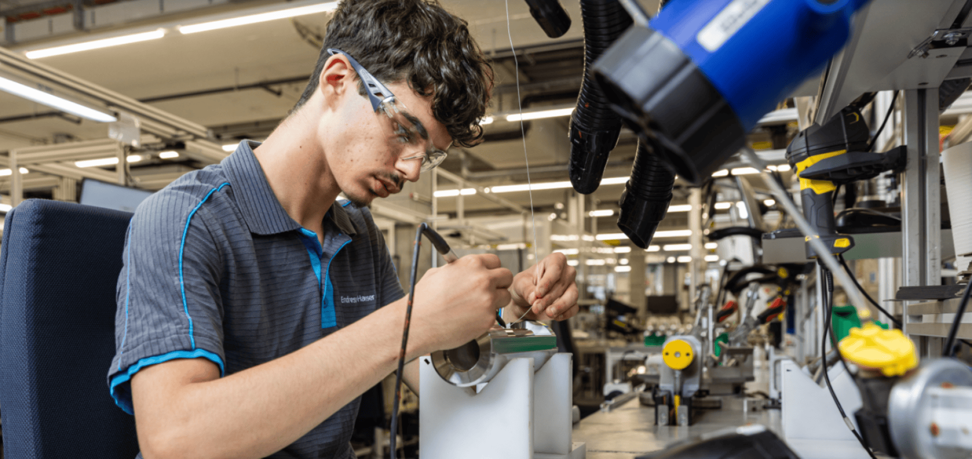 Ein junger Mann mit Schutzbrille arbeitet an einem Arbeitsplatz in einem modernen Industrielabor mit einem Lötgerät an elektronischen Bauteilen.