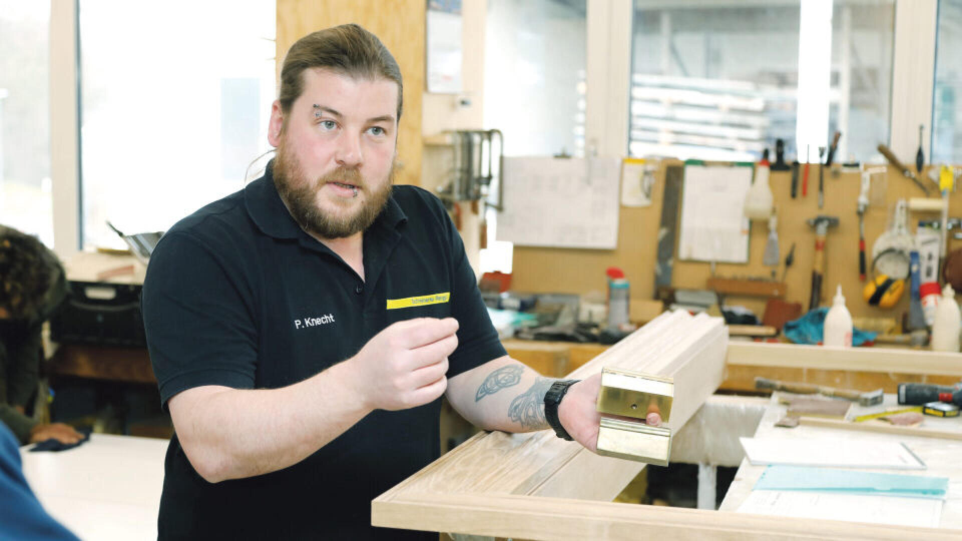 Ein Mann mit langen Haaren und Bart, der ein schwarzes Hemd trägt, erklärt etwas, während er ein Stück Holz und ein Metallwerkzeug in einer Werkstatt voller Werkzeuge und Papiere hält.