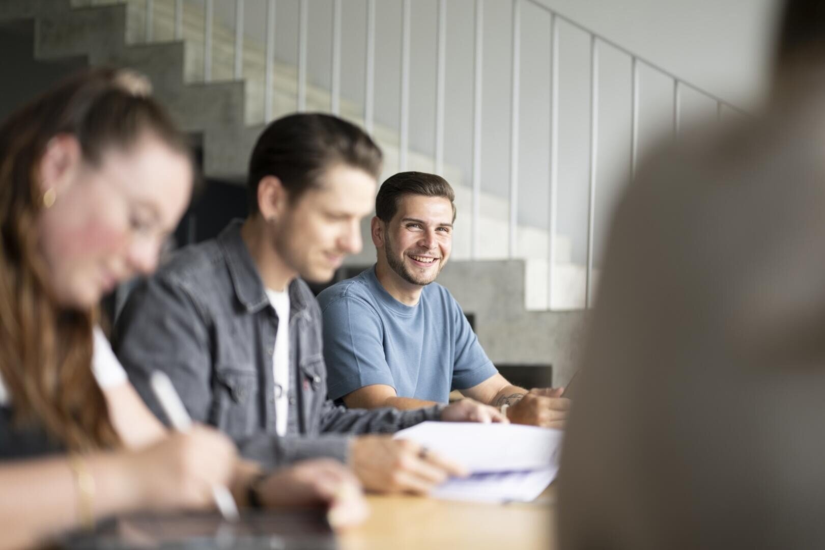 Eine Gruppe von Menschen sitzt an einem Tisch und schreibt oder arbeitet. Ein Mann in einem blauen Hemd steht im Fokus, lächelt und blickt in die Kamera, während andere im Vordergrund unscharf erscheinen. Im Hintergrund ist eine Treppe zu sehen.
