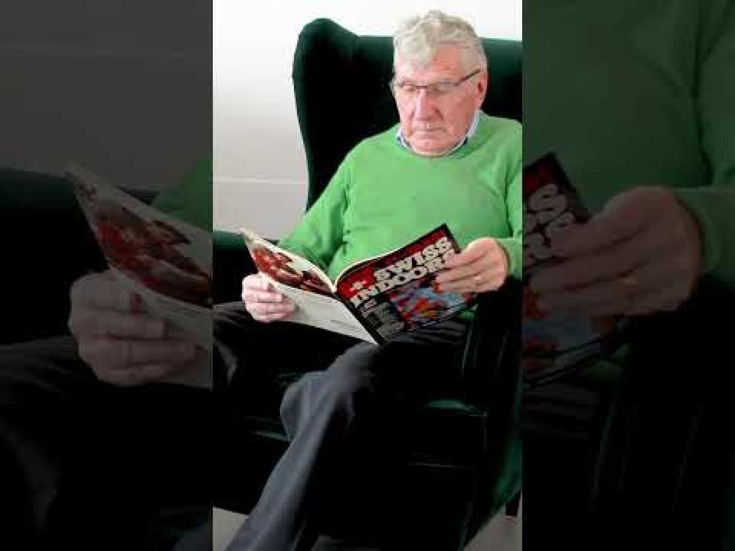 Ein älterer Mann mit grauem Haar und Brille, der einen hellgrünen Pullover trägt, sitzt in einem dunklen Stuhl und liest eine Zeitschrift mit dem Titel FourFourTwo. Er wirkt ruhig und konzentriert.