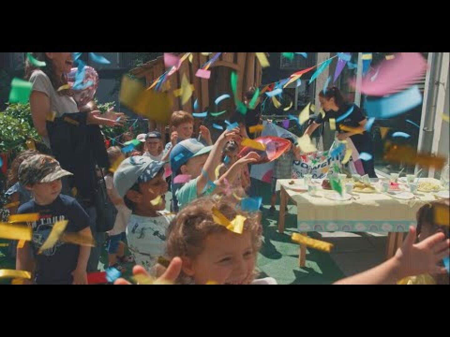 Kinder feiern auf einer Party im Freien, lächeln und spielen, während um sie herum buntes Konfetti fällt. Im Hintergrund steht ein Tisch mit Essen und Dekoration, und die Erwachsenen schauen zu. Die Szene ist lebhaft und festlich.