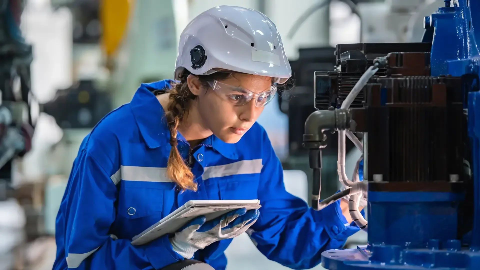 Eine Frau in einem blauen Overall, mit Schutzbrille und weißem Helm inspiziert Maschinen, während sie ein Tablet in einer Industrieumgebung hält.