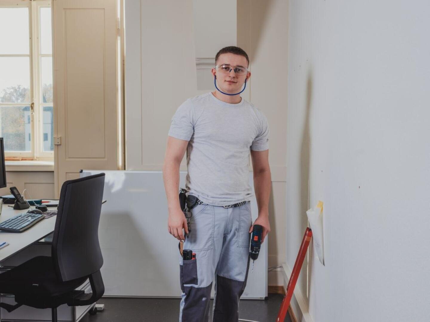 Ein Techniker, der eine Schutzbrille und eine Arbeitshose trägt, steht in einem Büro und hält einen Akku-Bohrer in der Hand.