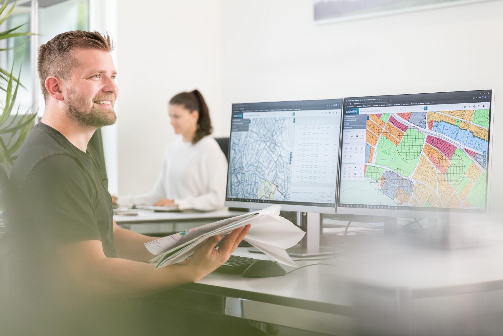 Ein lächelnder Mann hält Unterlagen in der Hand und sitzt an einem Schreibtisch, auf dem zwei Monitore stehen, auf denen Karten zu sehen sind. Im Hintergrund arbeitet eine Frau an einem Computer in einem hellen, modernen Büro.