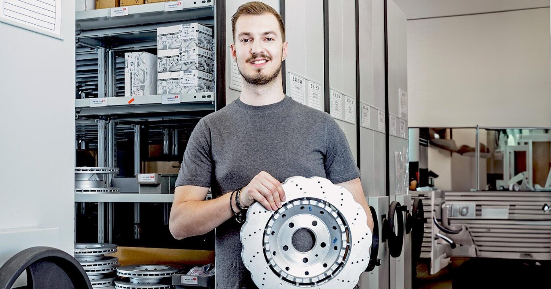 Ein Mann in einem grauen T-Shirt hält eine große, runde Metallbremsscheibe vor den Regalen einer Industriehalle in die Kamera.