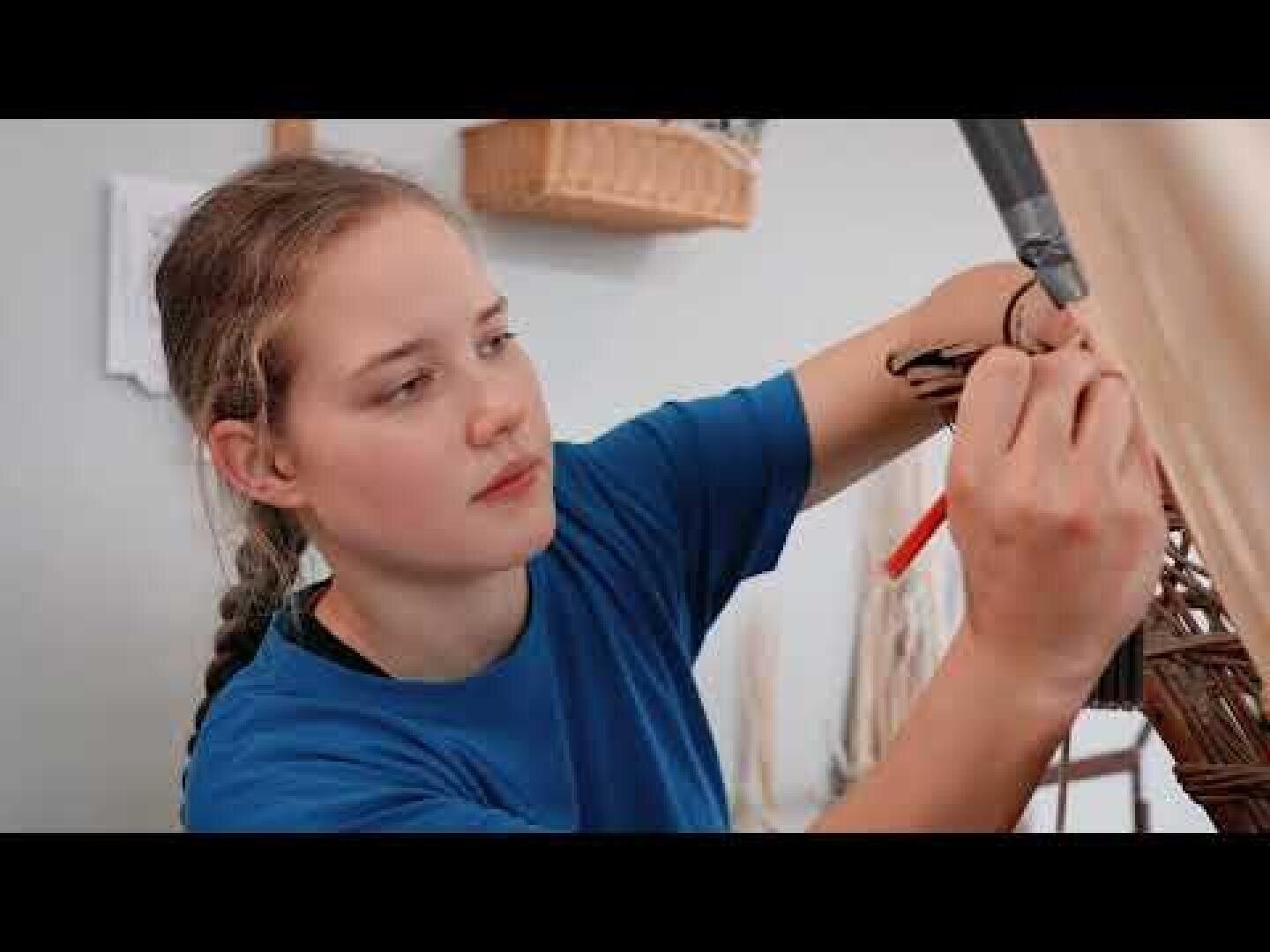 Eine junge Frau in einem blauen Hemd arbeitet konzentriert an einem handwerklichen Projekt, hält Werkzeuge in der Hand und webt oder fügt Materialien mit der Hand im Haus zusammen.
