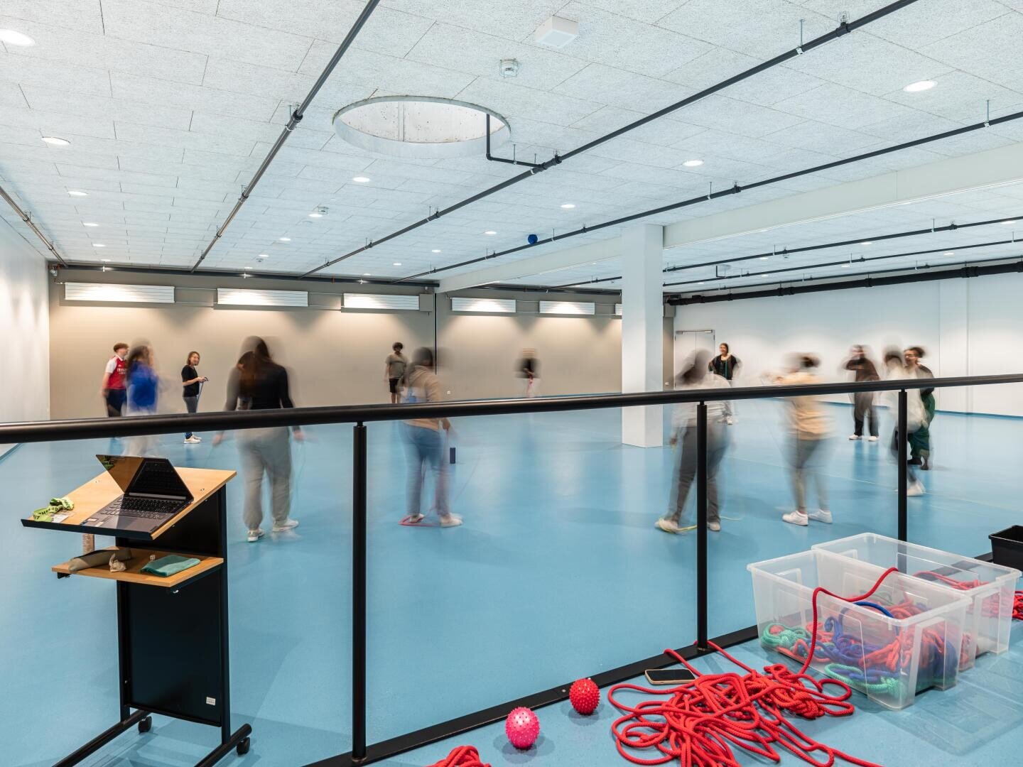 Eine geräumige Sporthalle, in der sich Menschen bewegen und herumlaufen. Im Vordergrund stehen Kisten mit Sportgeräten, ein kleiner Tisch mit einem Laptop und ein Geländer, das den Bereich von der Kamera trennt.