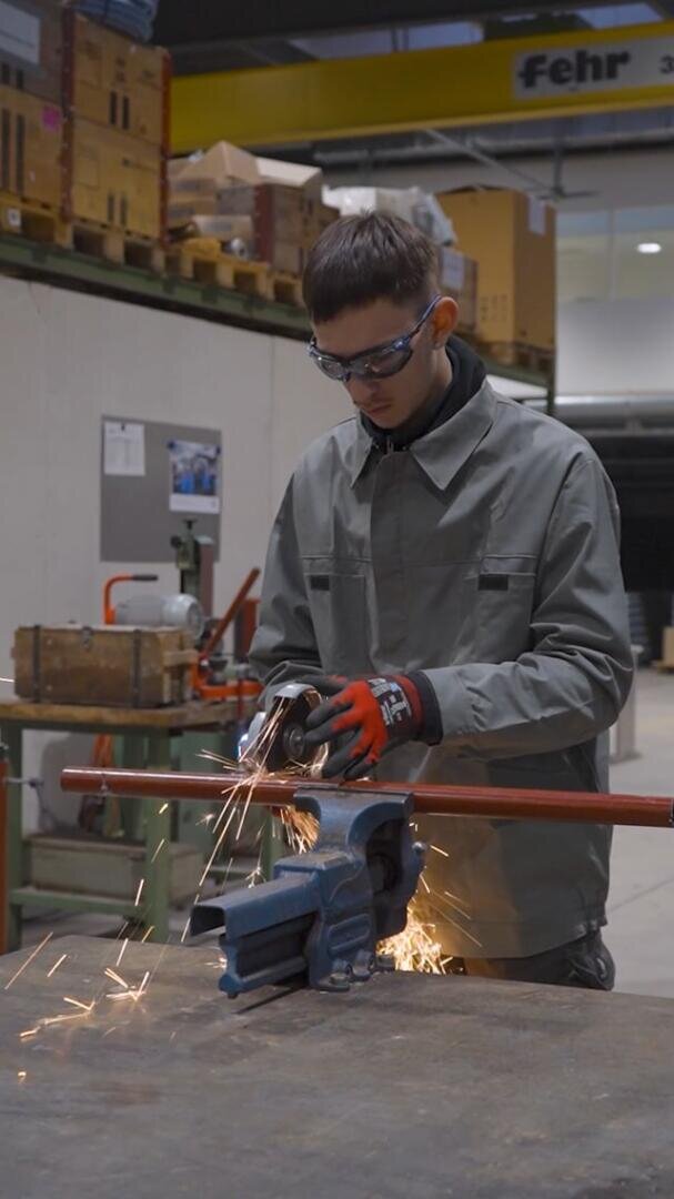 Ein Arbeiter, der eine Schutzbrille trägt, schleift ein Metallrohr, das in einem Schraubstock eingespannt ist, wobei Funken entstehen.
