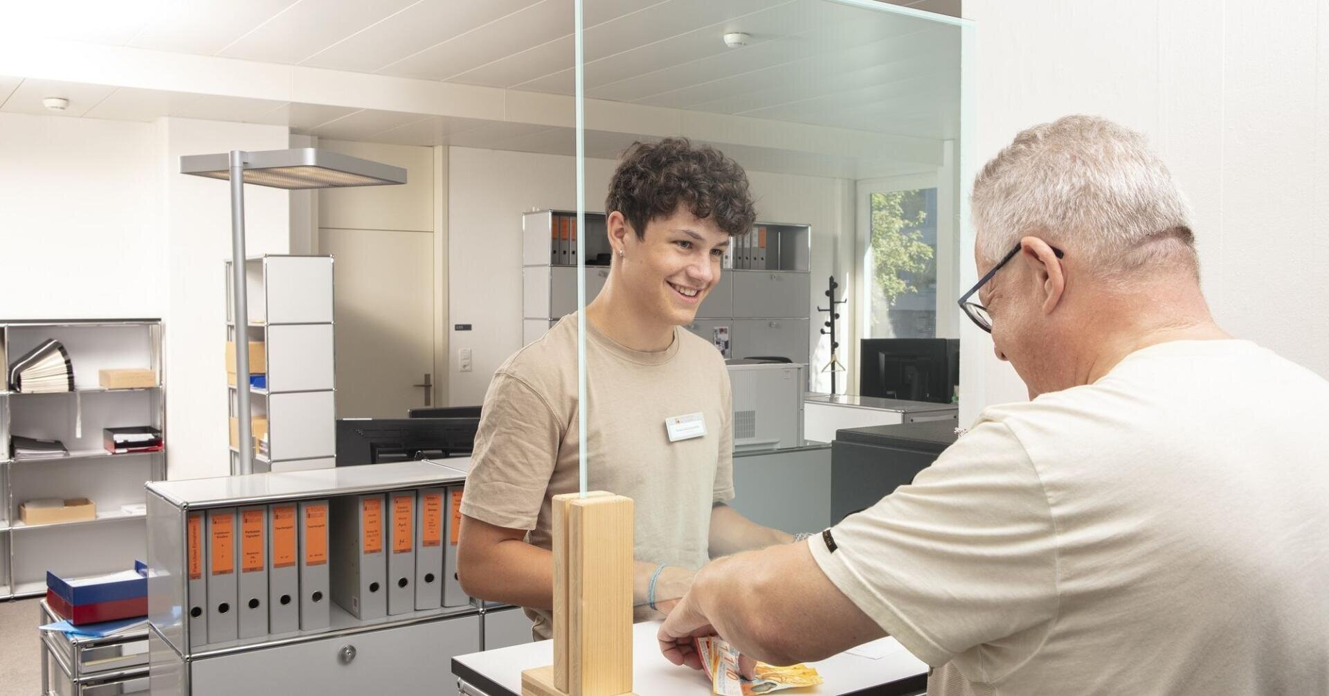 Ein junger Mann in einem beigen T-Shirt und mit einem Namensschild lächelt einen Kunden durch eine Glaswand hindurch in einem Büro an.