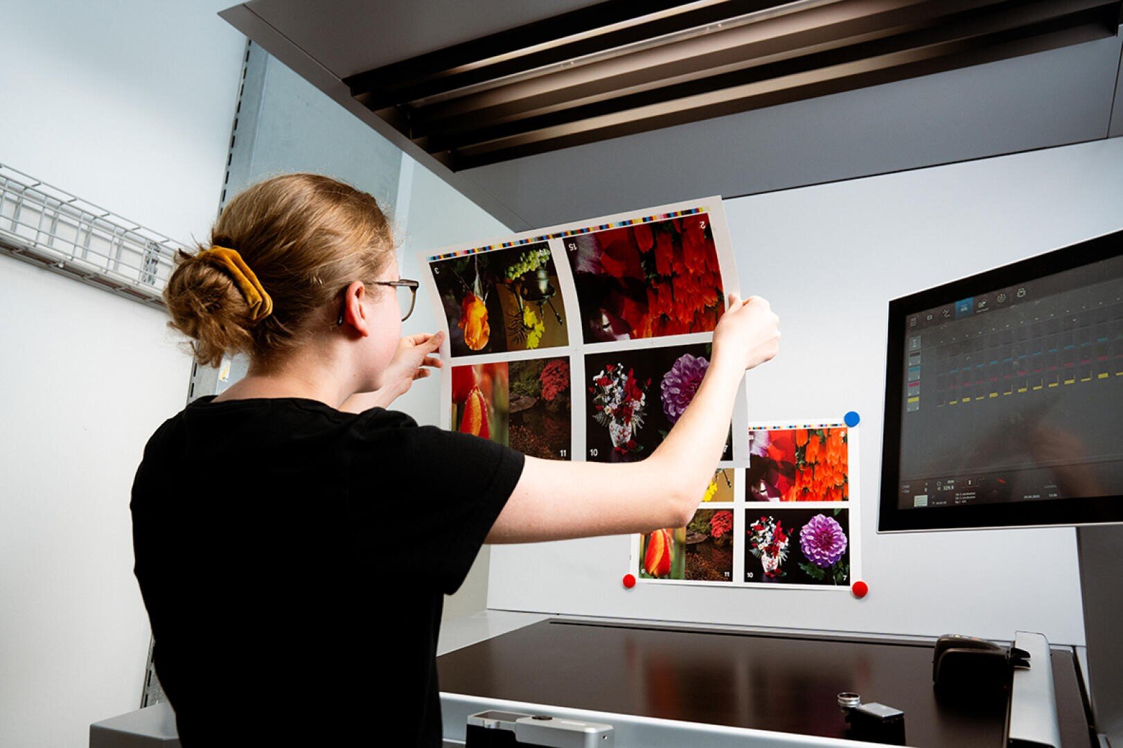 Eine Person begutachtet in einem professionellen Druck- oder Designstudio unter heller Beleuchtung in der Nähe eines Computermonitors gedruckte Blätter mit farbenfrohen Blumenfotos.