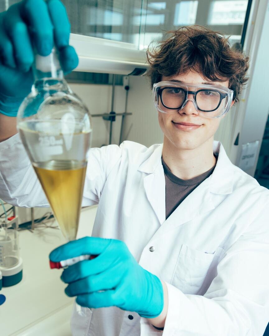 Ein junger Mensch in einem Laborkittel und mit Handschuhen hält einen Glas-Trenntrichter mit gelber Flüssigkeit in der Hand, steht in einem Labor und lächelt in die Kamera.