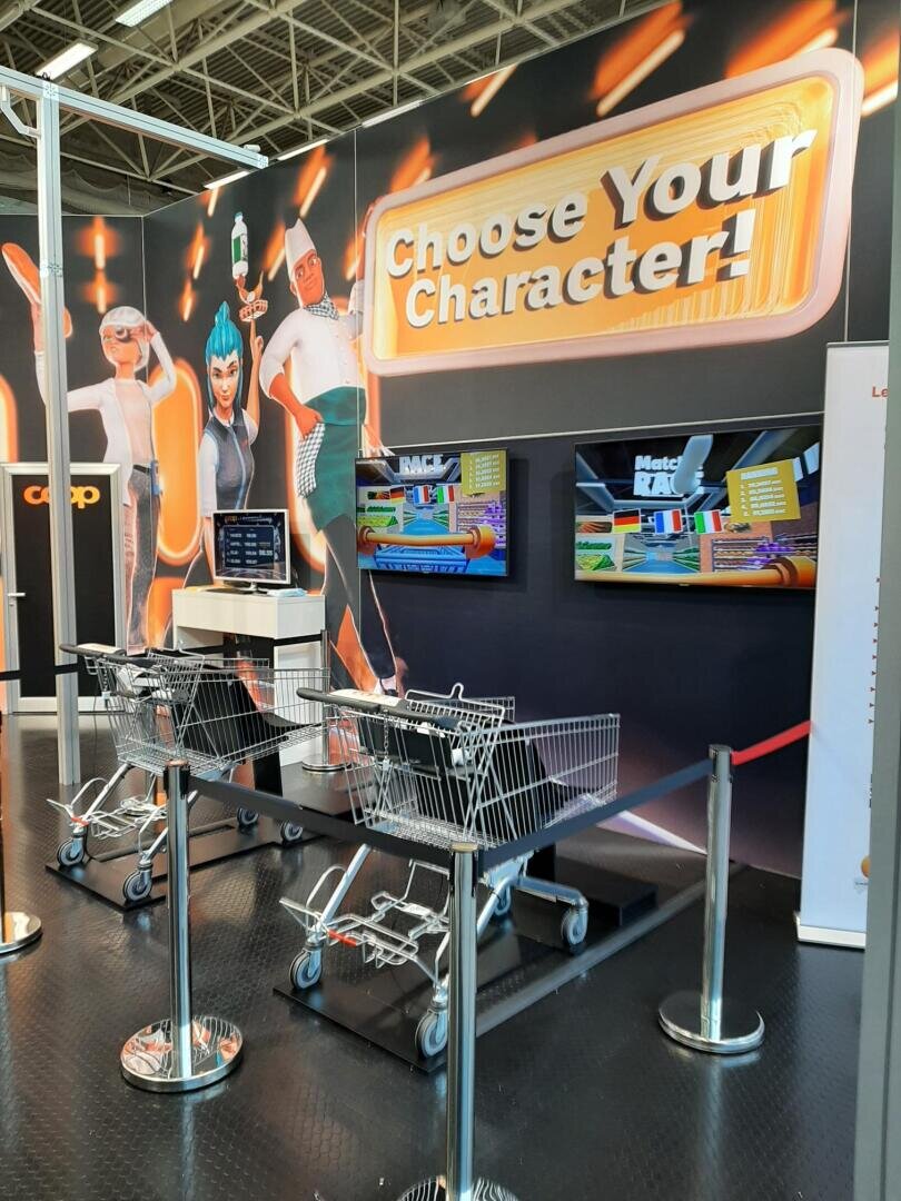 Drei leere Einkaufswagen sind vor Bildschirmen und einem großen Schild mit der Aufschrift "Choose Your Character!" aufgereiht. An der Rückwand sind animierte Figuren zu sehen, die an ein Videospiel in einem Geschäft erinnern.