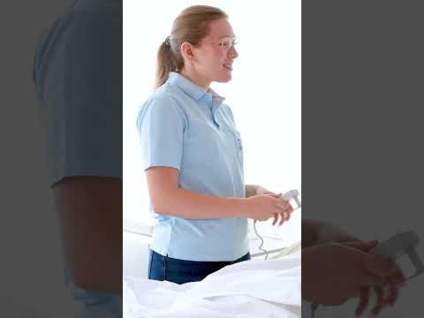 Eine Frau mit Brille und hellblauem Poloshirt steht in einem Haus und hält ein medizinisches Gerät in den Händen. Sie scheint sich in einer Klinik oder im Gesundheitswesen zu befinden.