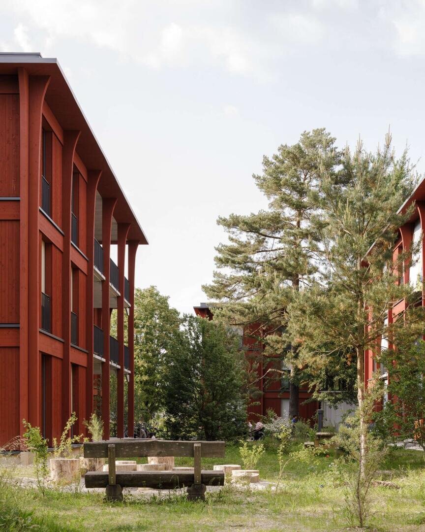 Ein Innenhof mit Rasen und Bäumen ist von modernen roten Wohnhäusern aus Holz umgeben. In der Mitte steht eine Bank aus Stein und Holz, und hohe Kiefern verleihen der Szenerie einen grünen Rahmen.