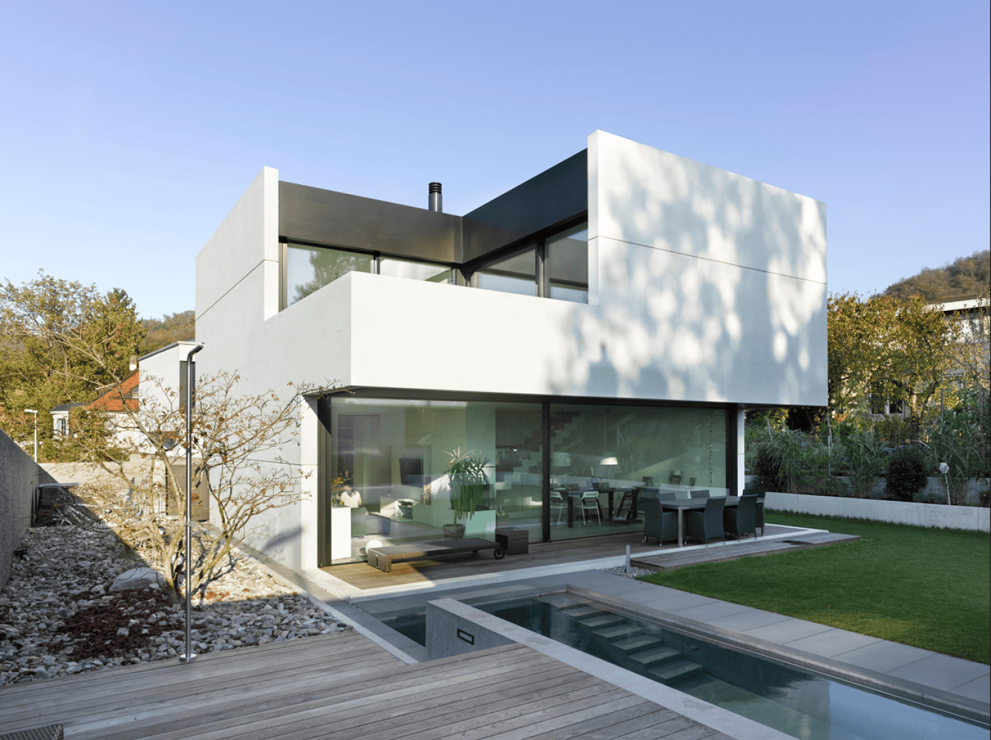 Ein modernes zweistöckiges Haus mit großen Glasfenstern, weißen Außenwänden, einem Essbereich im Freien, einem Pool und einer Holzterrasse, umgeben von viel Grün an einem sonnigen Tag.