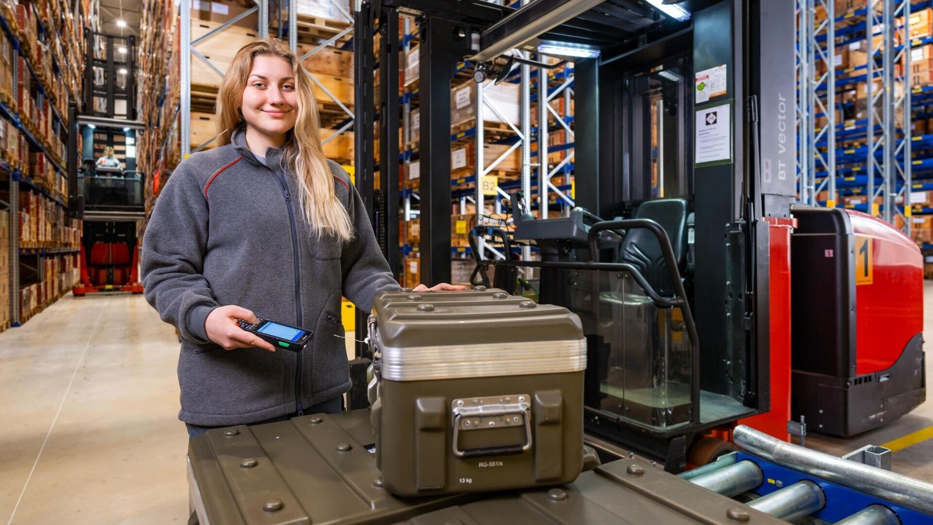 Eine Frau steht in einem Lagerhaus neben einem robusten Metallkoffer auf einem Wagen und hält einen Barcode-Scanner in der Hand. Im Hintergrund sind mit Kartons gefüllte Regale und ein roter Gabelstapler zu sehen.