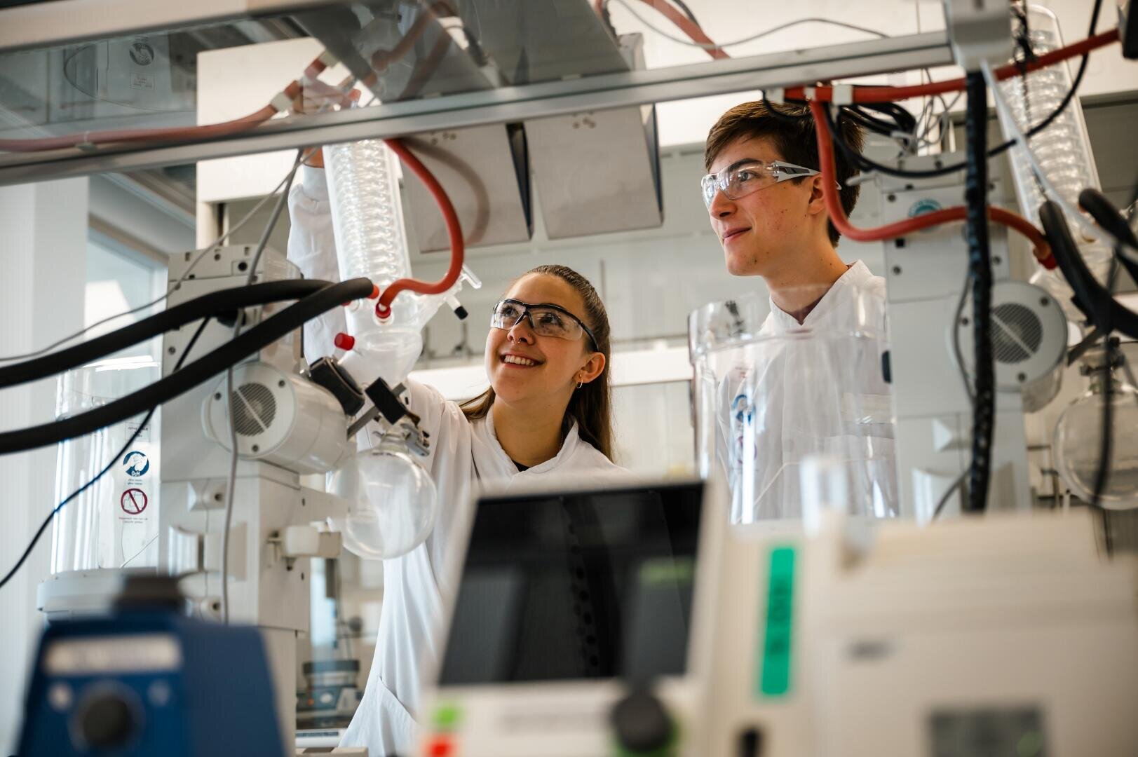 Zwei Forscher in Laborkitteln und mit Schutzbrillen arbeiten in einem modernen Labor mit wissenschaftlichen Geräten und Glasgeräten, lächeln und konzentrieren sich auf ihr Experiment.