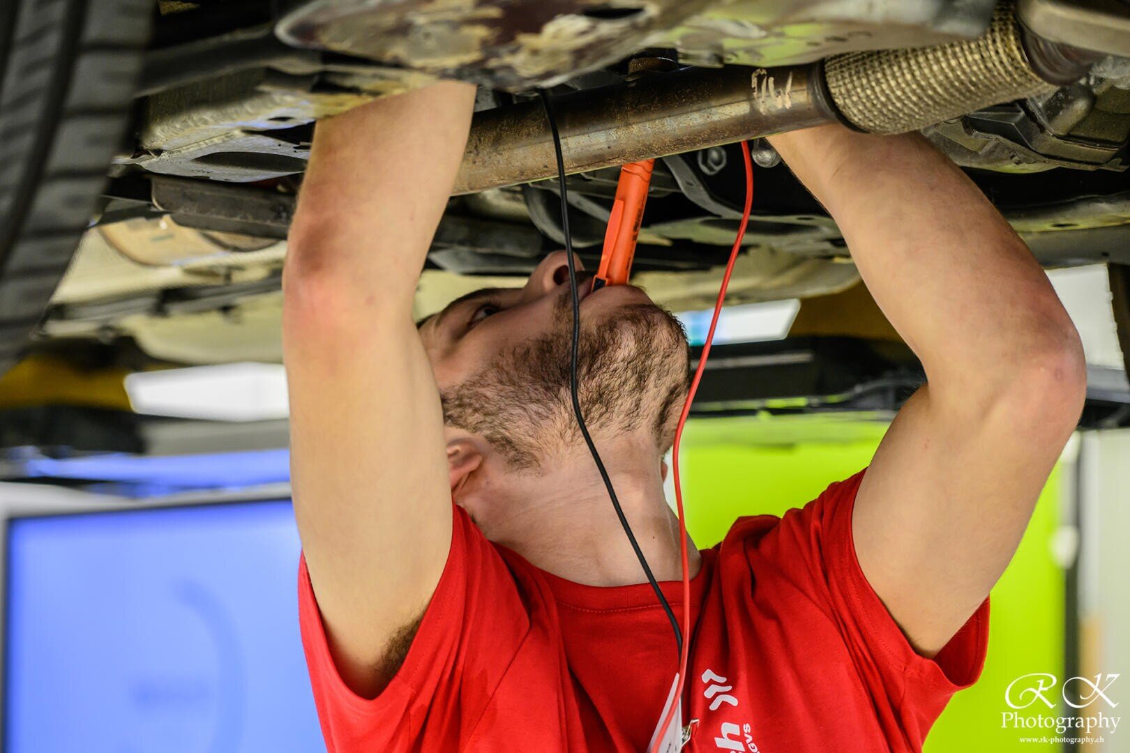 Eine Person in einem roten Hemd untersucht mit einem Diagnosegerät die Unterseite eines auf einer Hebebühne angehobenen Autos, hält dabei Kabel fest und betrachtet die Fahrzeugkomponenten genau.