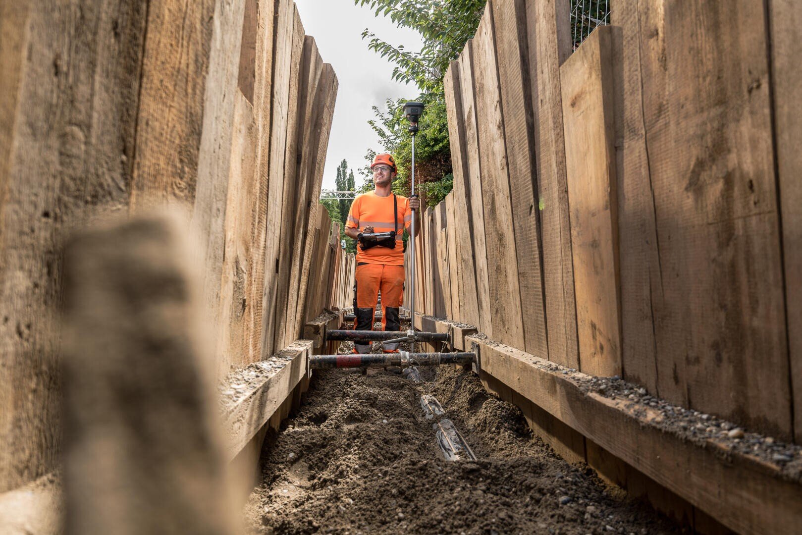 Ein Bauarbeiter in orangefarbener Schutzkleidung steht in einem tiefen, mit Holzbrettern ausgekleideten Graben, umgeben von Erde und Rohren, im Hintergrund sind Bäume zu sehen.