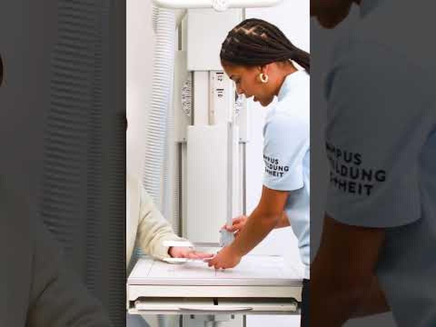 Ein Mitarbeiter des Gesundheitswesens hilft einem Patienten, indem er dessen Hand auf einen medizinischen Bildgebungstisch legt, wahrscheinlich für eine Röntgenaufnahme in einer klinischen Umgebung. Die Mitarbeiterin trägt eine hellblaue Uniform mit Text auf dem Ärmel.