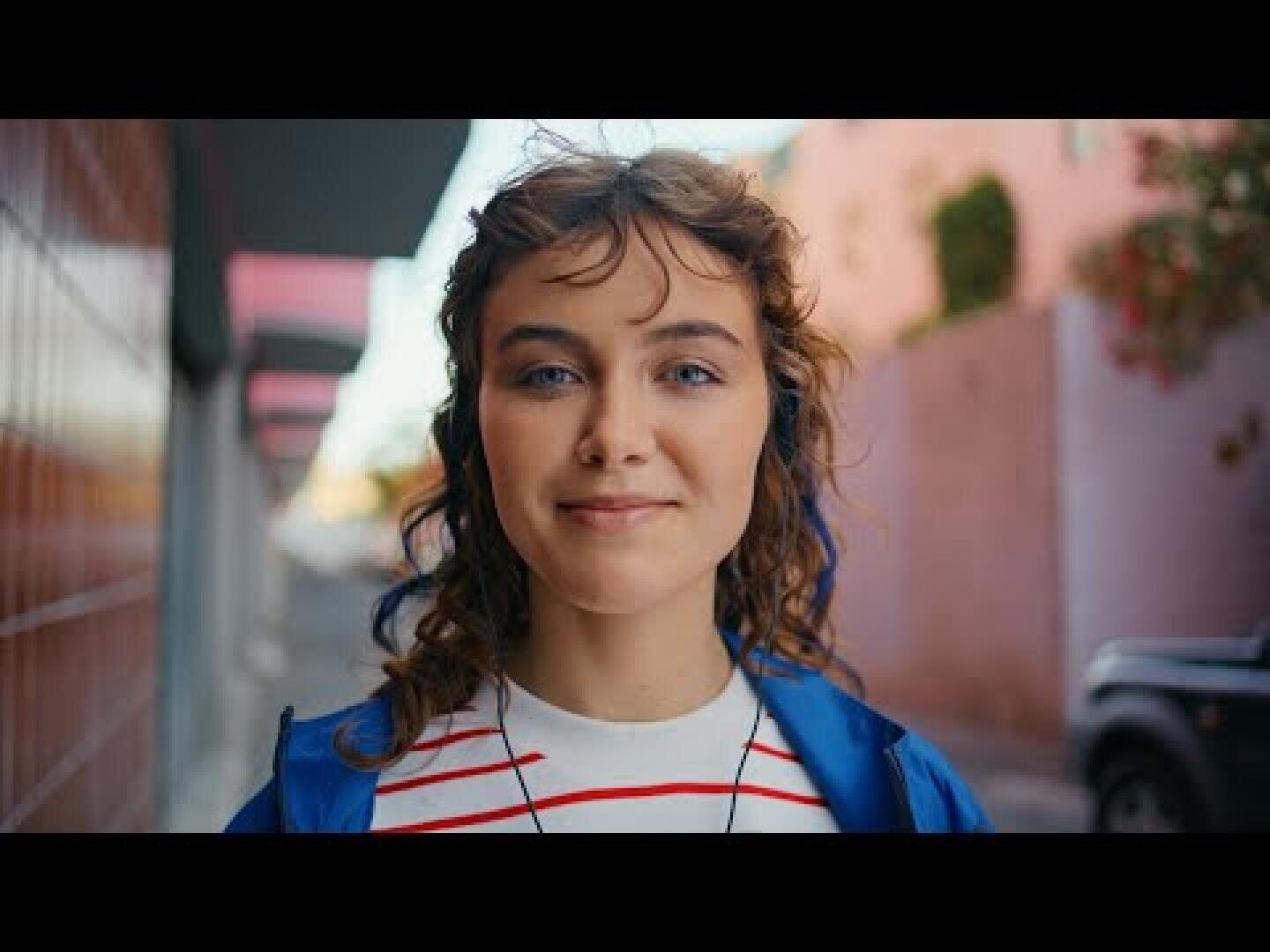 Eine junge Frau mit lockigem Haar lächelt ins Objektiv, während sie im Freien steht. Sie trägt ein gestreiftes Shirt und eine blaue Jacke; im Hintergrund ist eine verschwommene Stadtkulisse mit rosa Wänden und einem geparkten Auto zu sehen.