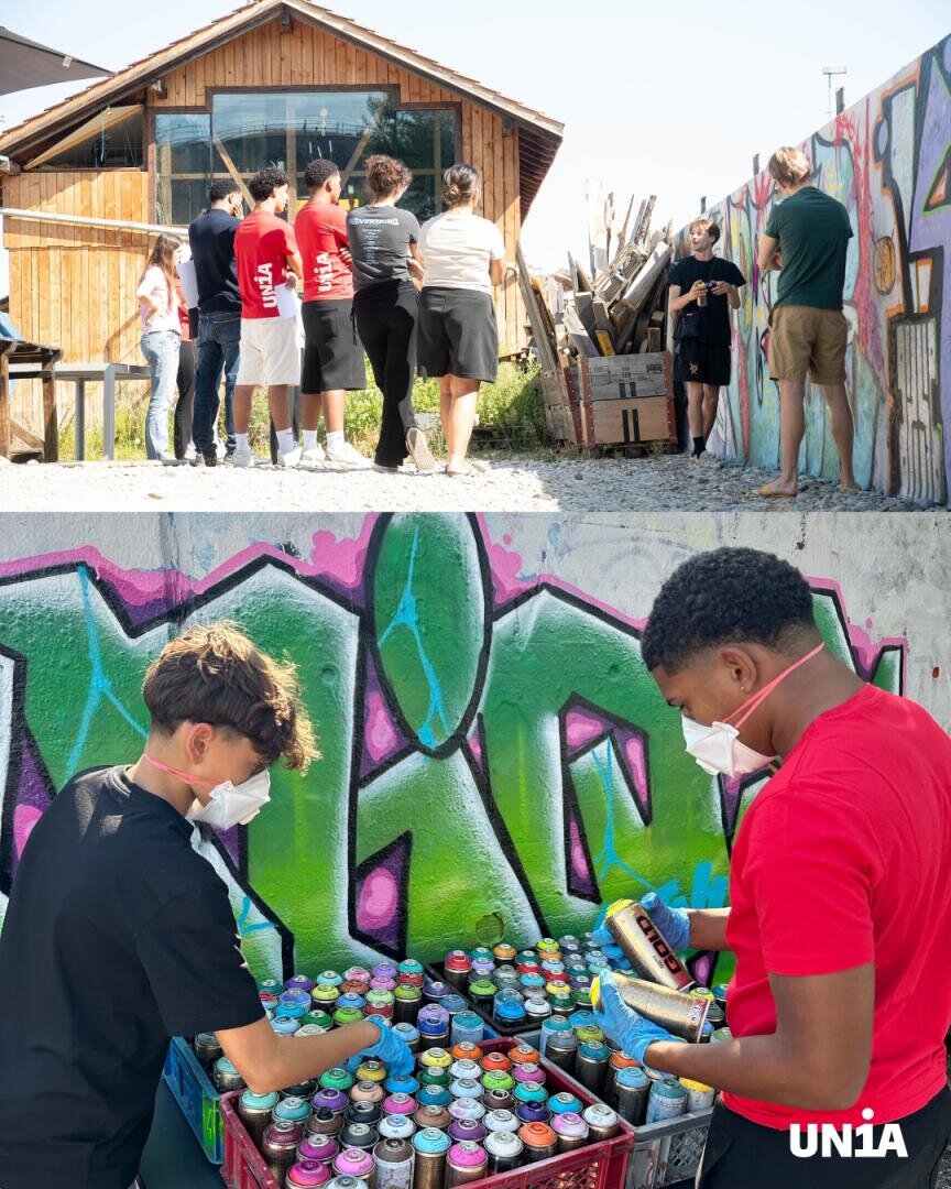 Zwei Teenager mit Masken suchen sich in der Nähe einer bunten Graffiti-Wand Sprühdosen aus, während sich im Hintergrund eine Gruppe von Menschen vor einem Holzgebäude versammelt. Unten rechts ist das UNIA-Logo zu sehen.