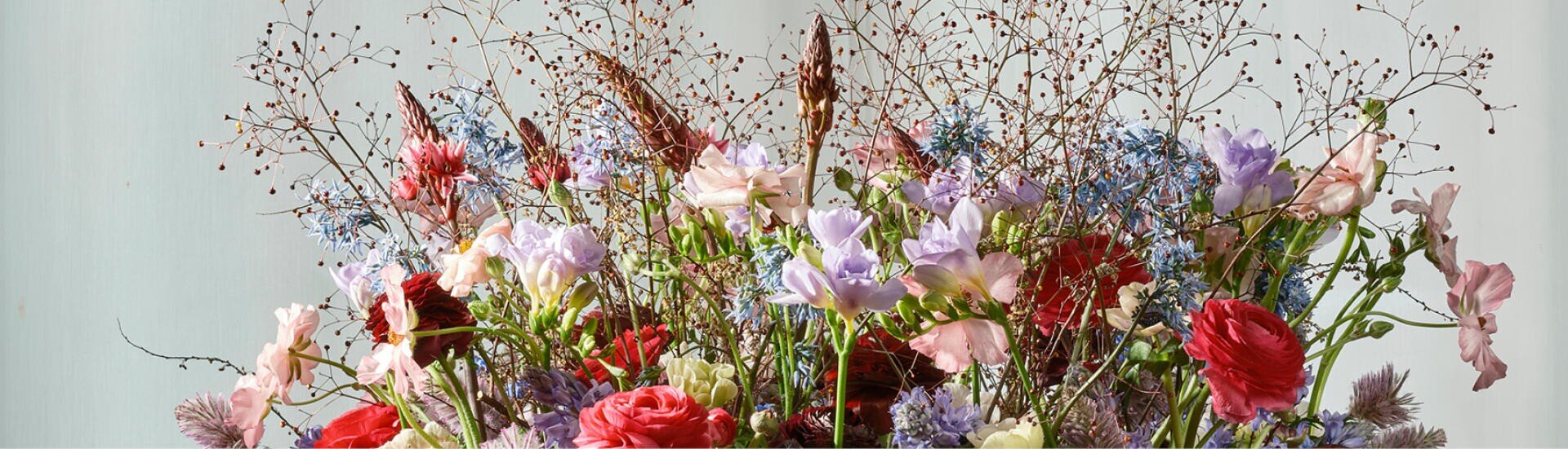 Ein farbenfrohes Arrangement aus verschiedenen Wildblumen, darunter rosa, rote, lavendelfarbene und blaue Blüten, ergänzt durch zarte braune Zweige und Grün, vor einem schlichten, hellen Hintergrund.