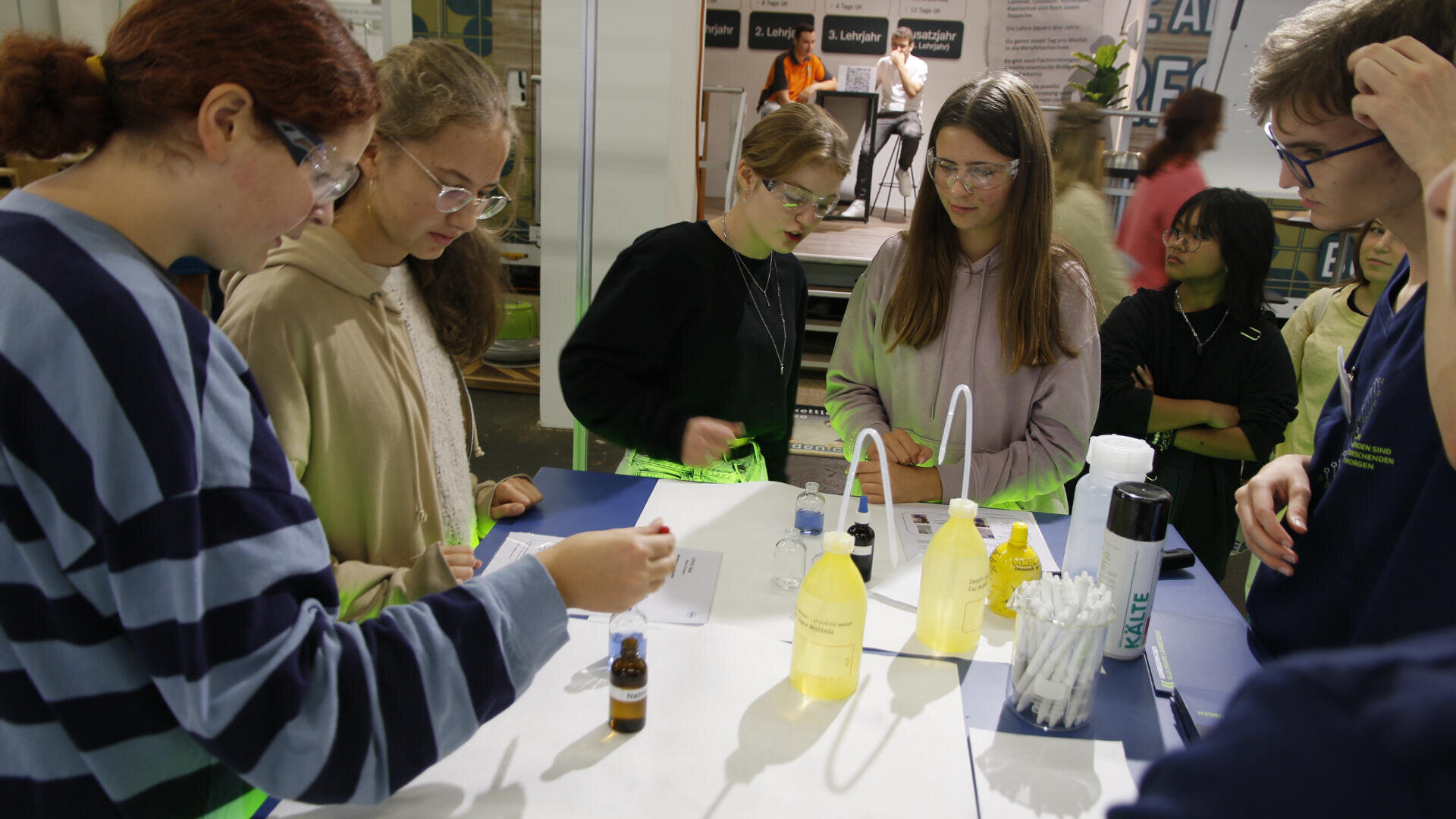 Fünf Schüler mit Schutzbrillen versammeln sich um einen Labortisch mit gelben Flüssigkeitsflaschen, Tropfern und Papier und führen ein wissenschaftliches Experiment durch. Ein Erwachsener und andere Personen sind im Hintergrund zu sehen.