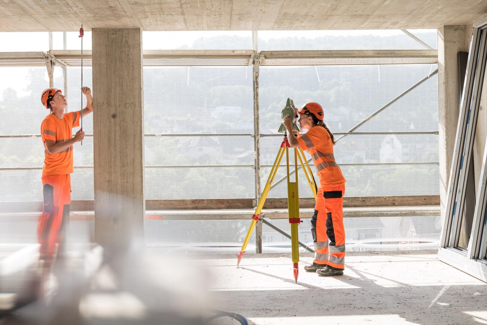 Zwei Bauarbeiter in orangefarbener Schutzkleidung verwenden Vermessungsgeräte und nehmen Messungen in einem im Bau befindlichen Gebäude vor, im Hintergrund sind Gerüste und große Fenster zu sehen.