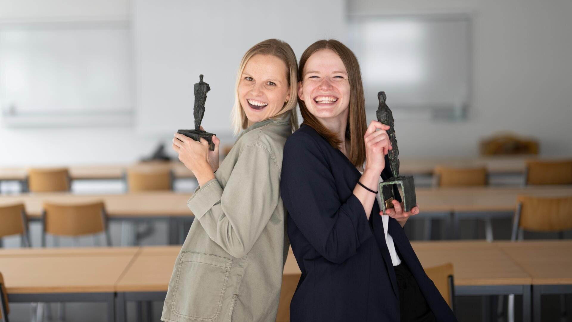 Zwei junge Frauen stehen Rücken an Rücken in einem Klassenzimmer, lächeln und halten kleine Trophäenstatuen. Im Hintergrund sind leere Tische und Stühle sowie ein Whiteboard zu sehen.