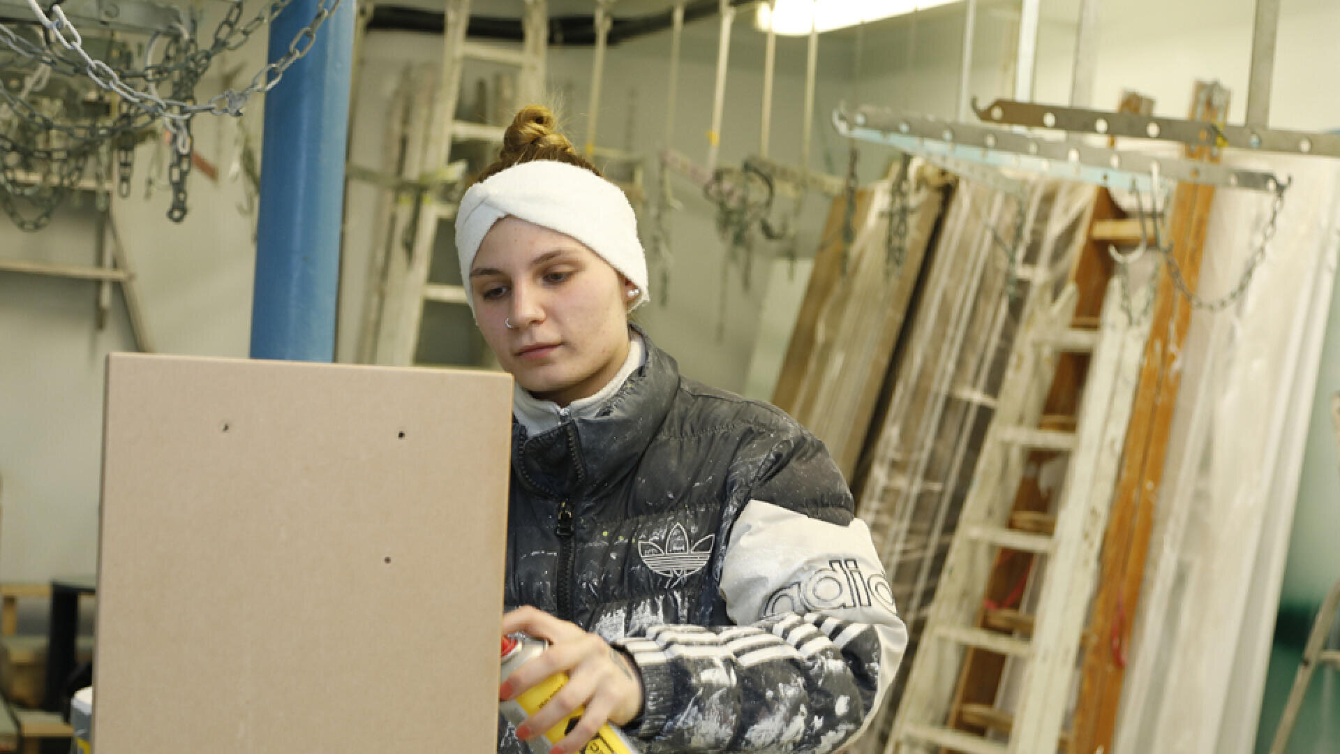 Eine Person, die ein weißes Stirnband und eine Adidas-Jacke trägt, arbeitet in einer Werkstatt an einer Holzplatte, umgeben von Leitern, Ketten und verschiedenen Baumaterialien.