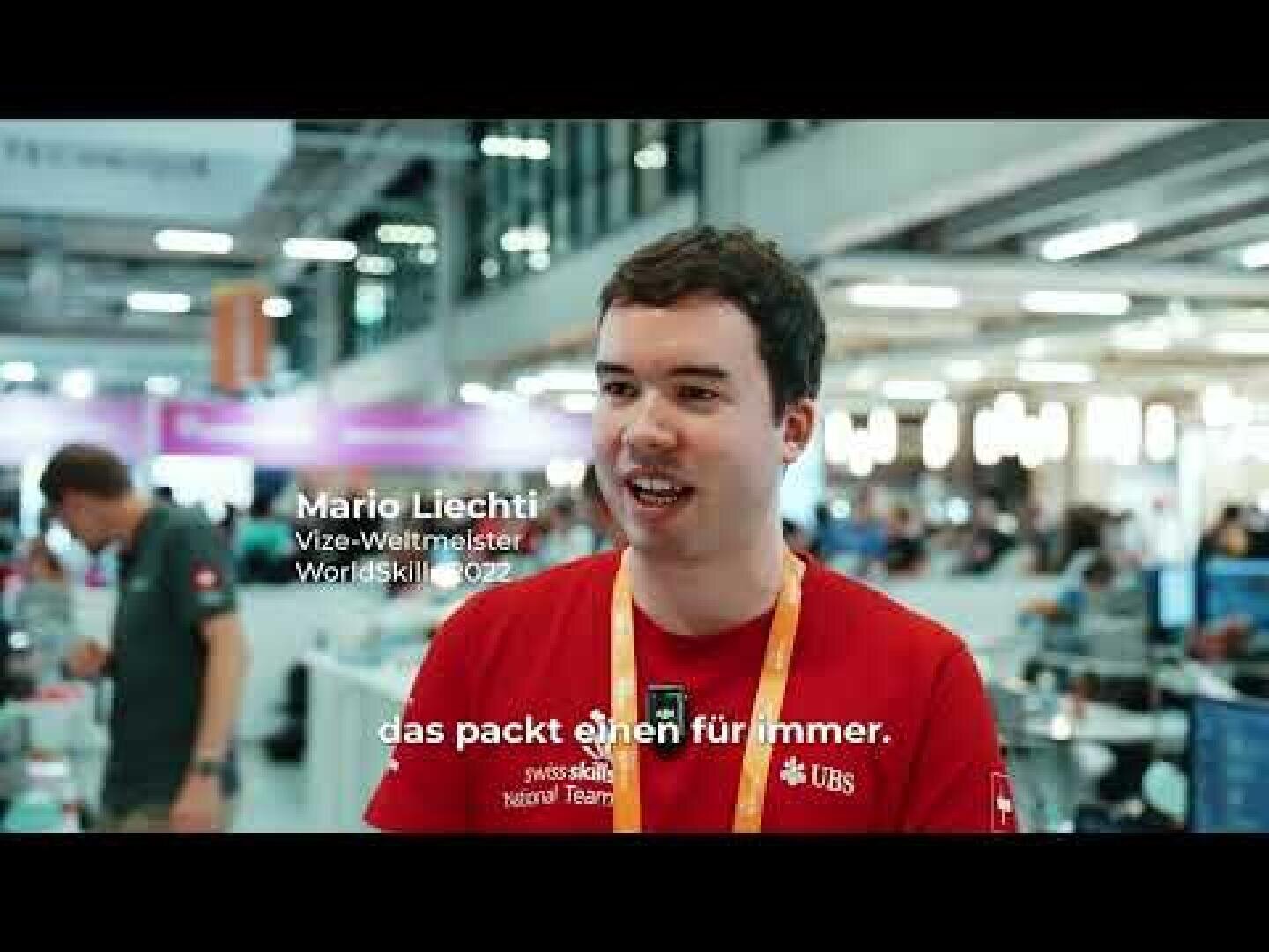 Ein junger Mann, der ein rotes Hemd mit UBS-Logo und ein gelbes Schlüsselband trägt, steht in einem Innenraum an einer belebten Veranstaltung. Der Text auf dem Bild lautet: "Mario Liechti, Vize-Weltmeister, WorldSkill 2022" und "das packt einen für immer.".