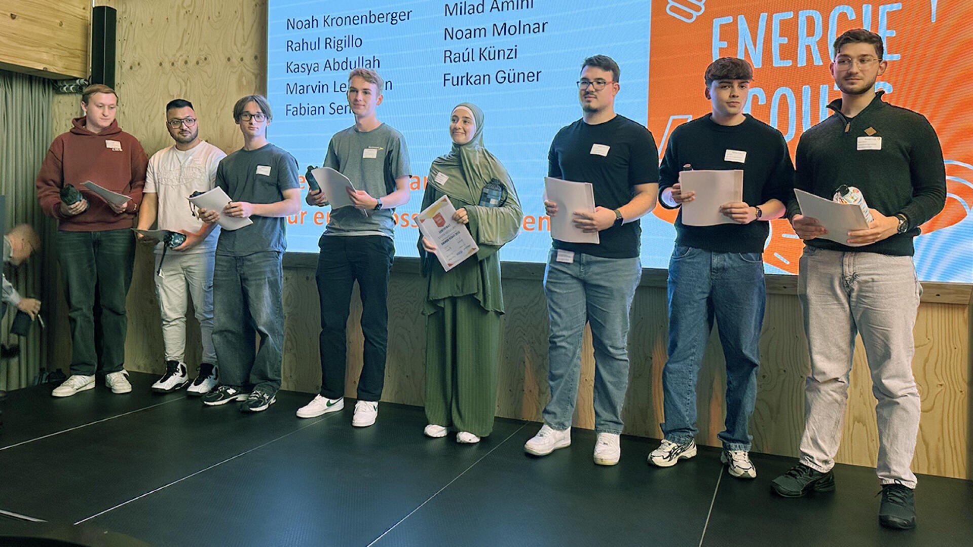 Acht Personen stehen in einer Reihe auf einer Bühne und halten Urkunden in der Hand. Hinter ihnen befindet sich ein Präsentationsbildschirm mit Namen und einer orangefarbenen Grafik. Sie scheinen an einer Preisverleihung oder Anerkennungsveranstaltung teilzunehmen.