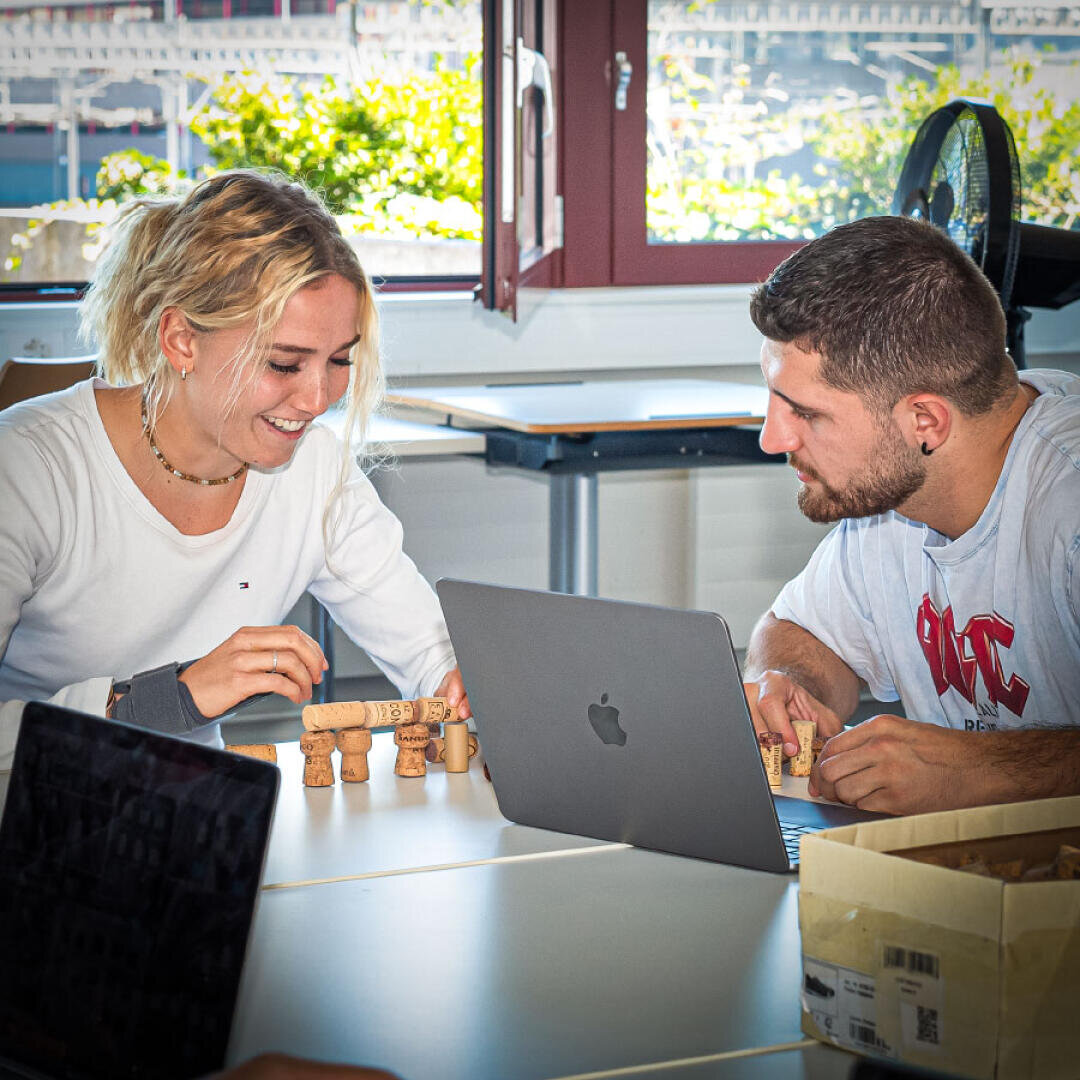 Zwei Menschen sitzen an einem Tisch mit einem Laptop, lächeln und spielen mit kleinen Korkfiguren in einem hellen Raum mit einem Fenster, vor dem grüne Pflanzen stehen.