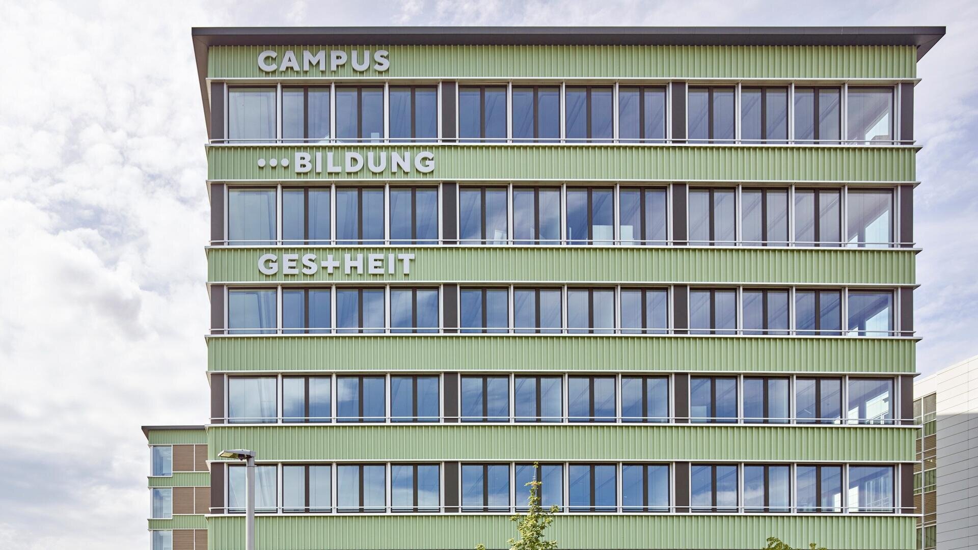 Ein modernes, umweltfreundliches Gebäude mit großen Fenstern, auf dessen oberen Stockwerken vor einem teilweise bewölkten Himmel die deutschen Wörter CAMPUS, … BILDUNG und GES+HEIT zu sehen sind.